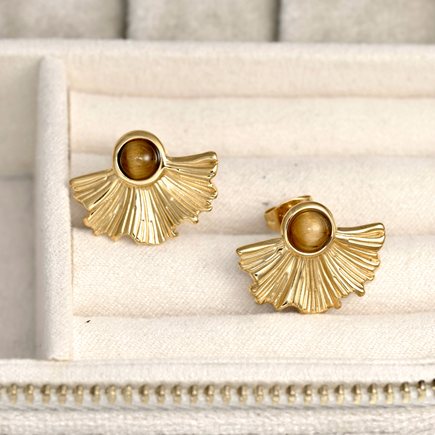 A pair of gold fan stud earrings with round brown tiger's eye stones, displayed on a cream-colored jewelry box.