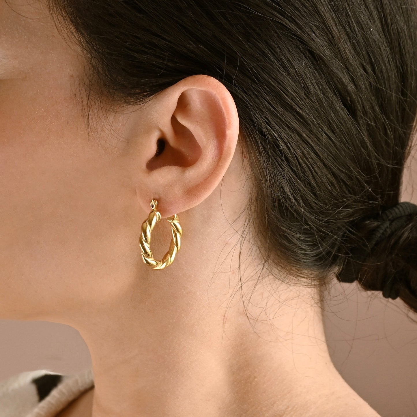 A close-up of a person's ear wearing a twisted gold hoop earring, with dark hair tied back in a low bun against a soft neutral background.