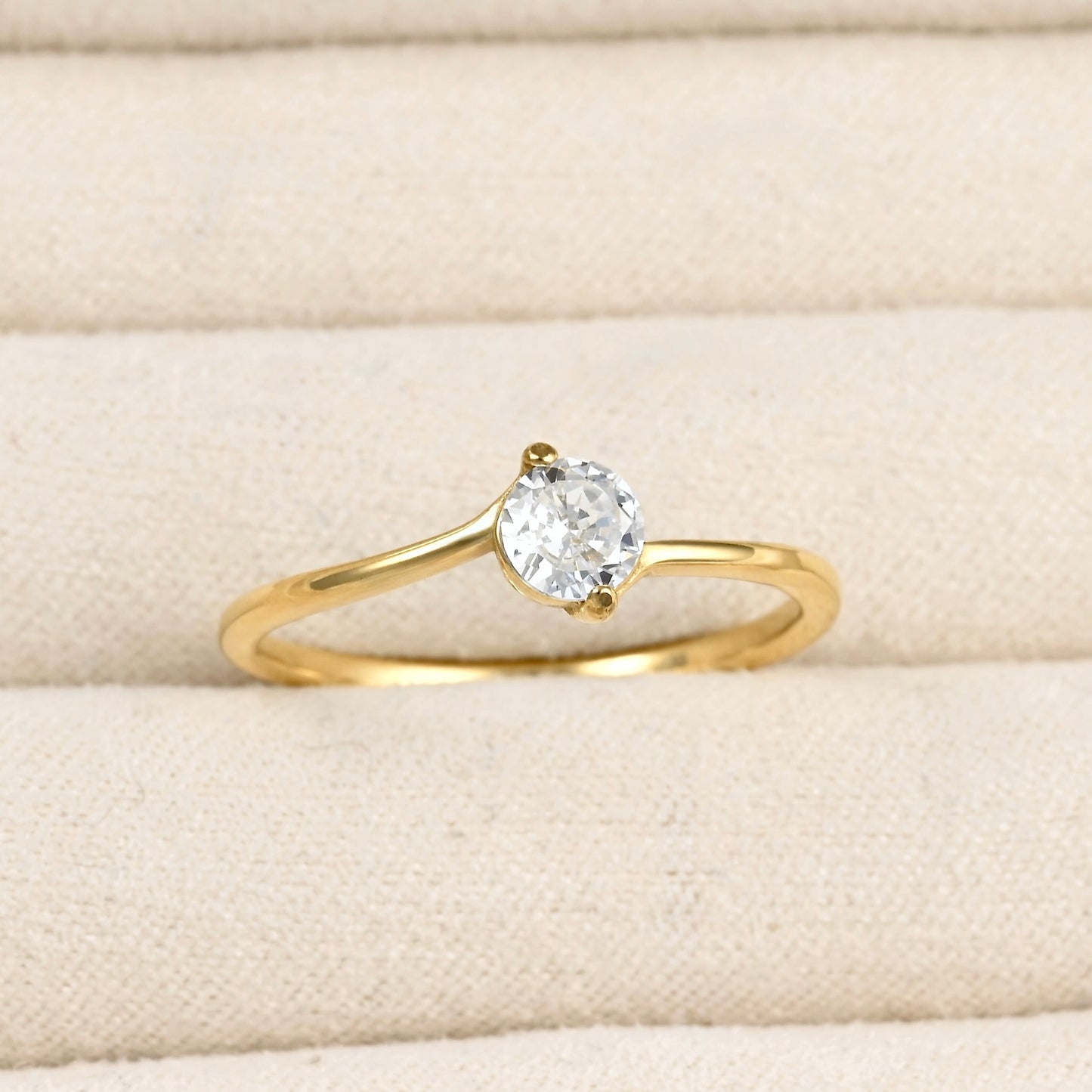 A yellow gold solitaire ring with a round-cut clear gemstone, displayed on a textured beige fabric ring box.