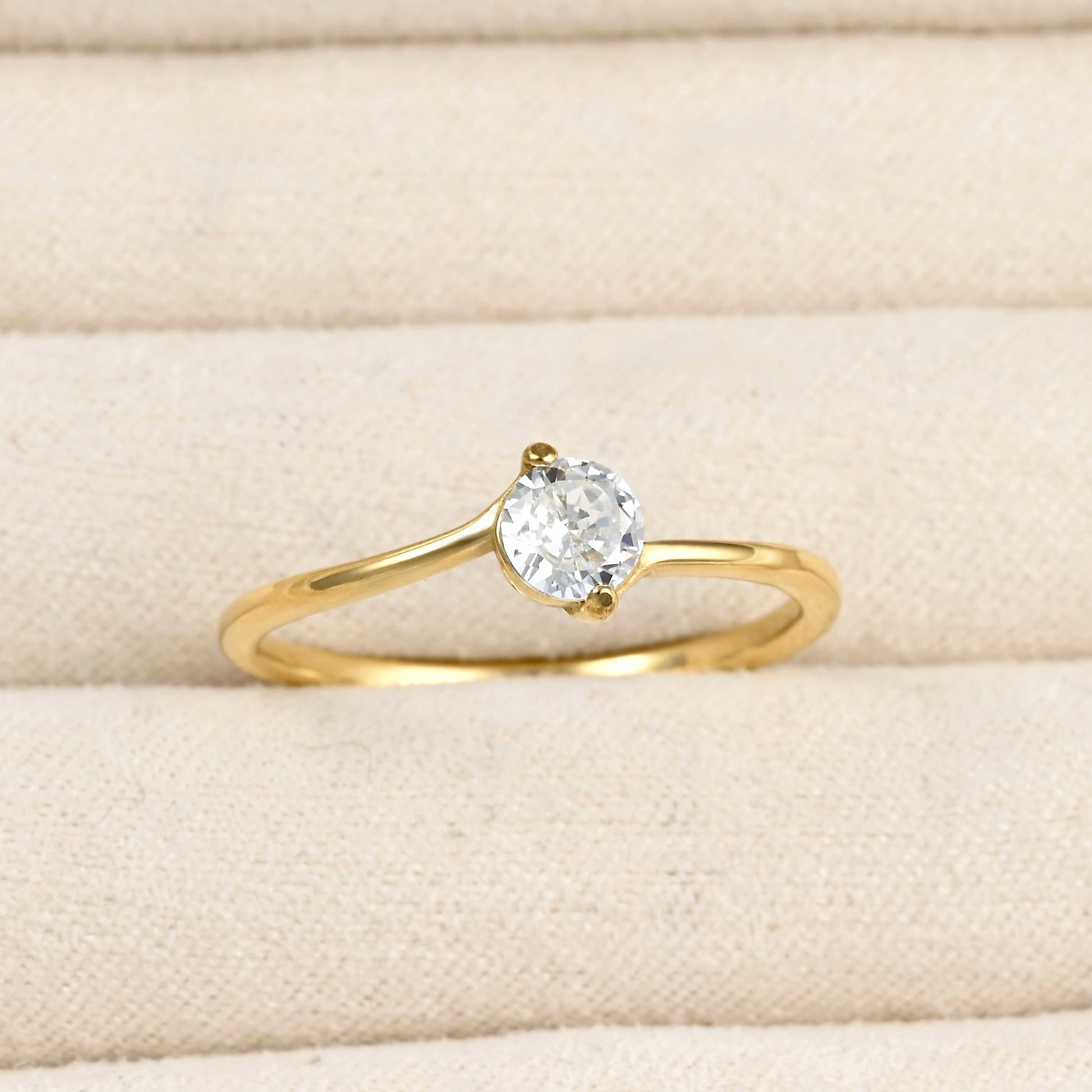 A yellow gold solitaire ring with a round-cut clear gemstone, displayed on a textured beige fabric ring box.