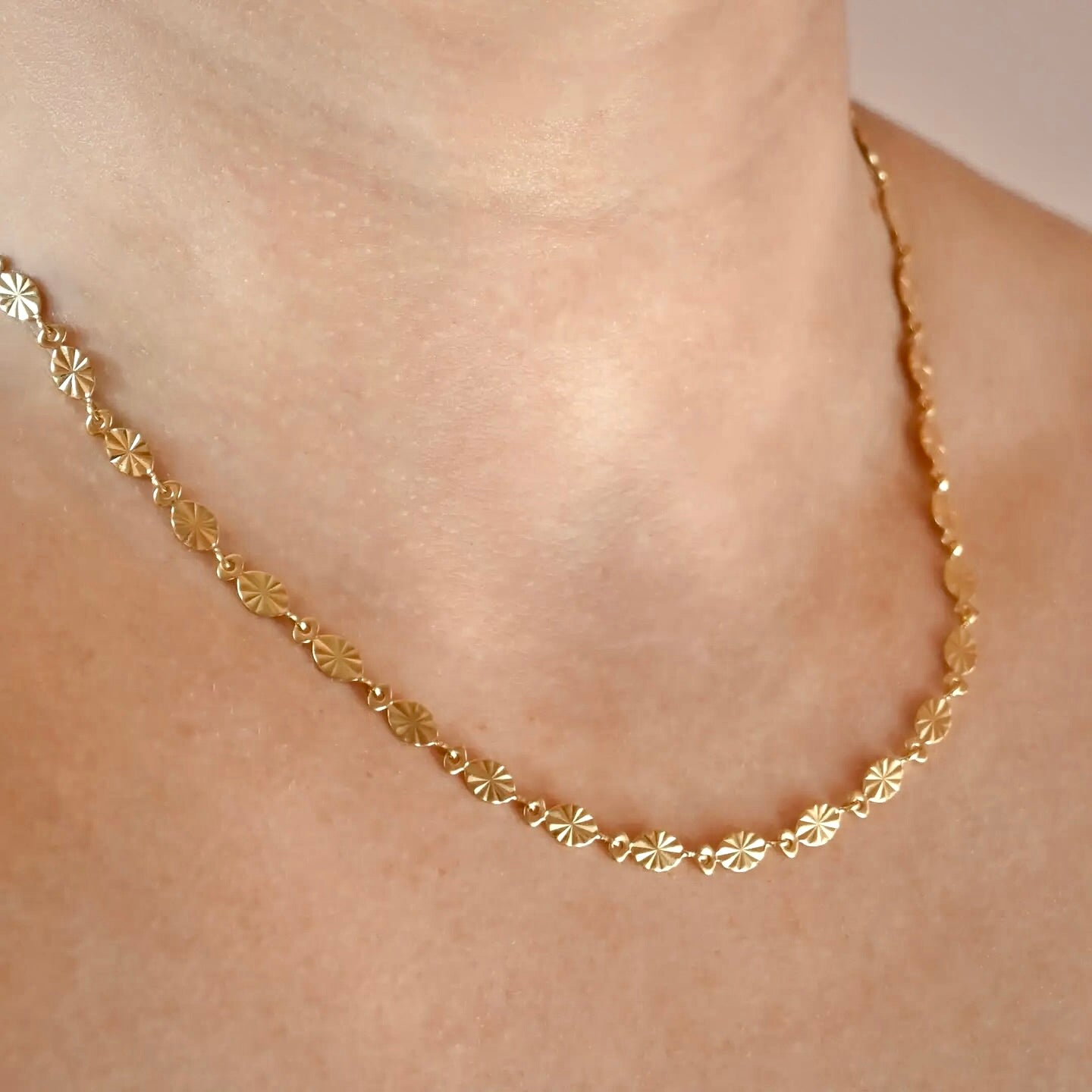 A delicate gold chain necklace with small, repeating sunburst links, shown worn on a person's neck against a soft beige background.