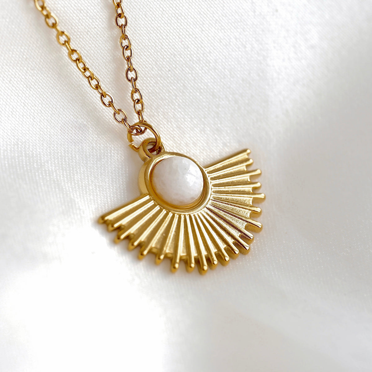 A gold pendant necklace featuring a sunburst fan design with a central white round moonstone, shown on a white textured fabric background.