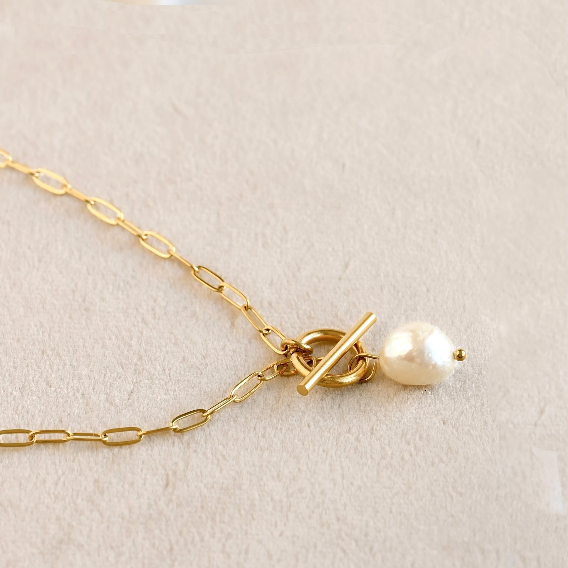 A close-up of a gold chain necklace with a toggle clasp and a small white pearl bead, displayed on a soft beige textured surface.
