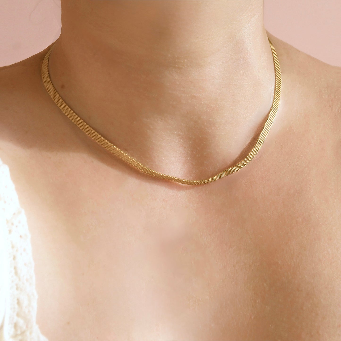 A close-up of a person's neck and upper chest wearing a delicate gold mesh chain necklace against a soft pink background.