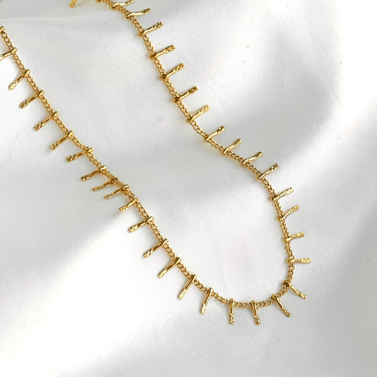 A gold chain necklace featuring small, textured vertical spikes along the front, displayed on a soft white fabric background.