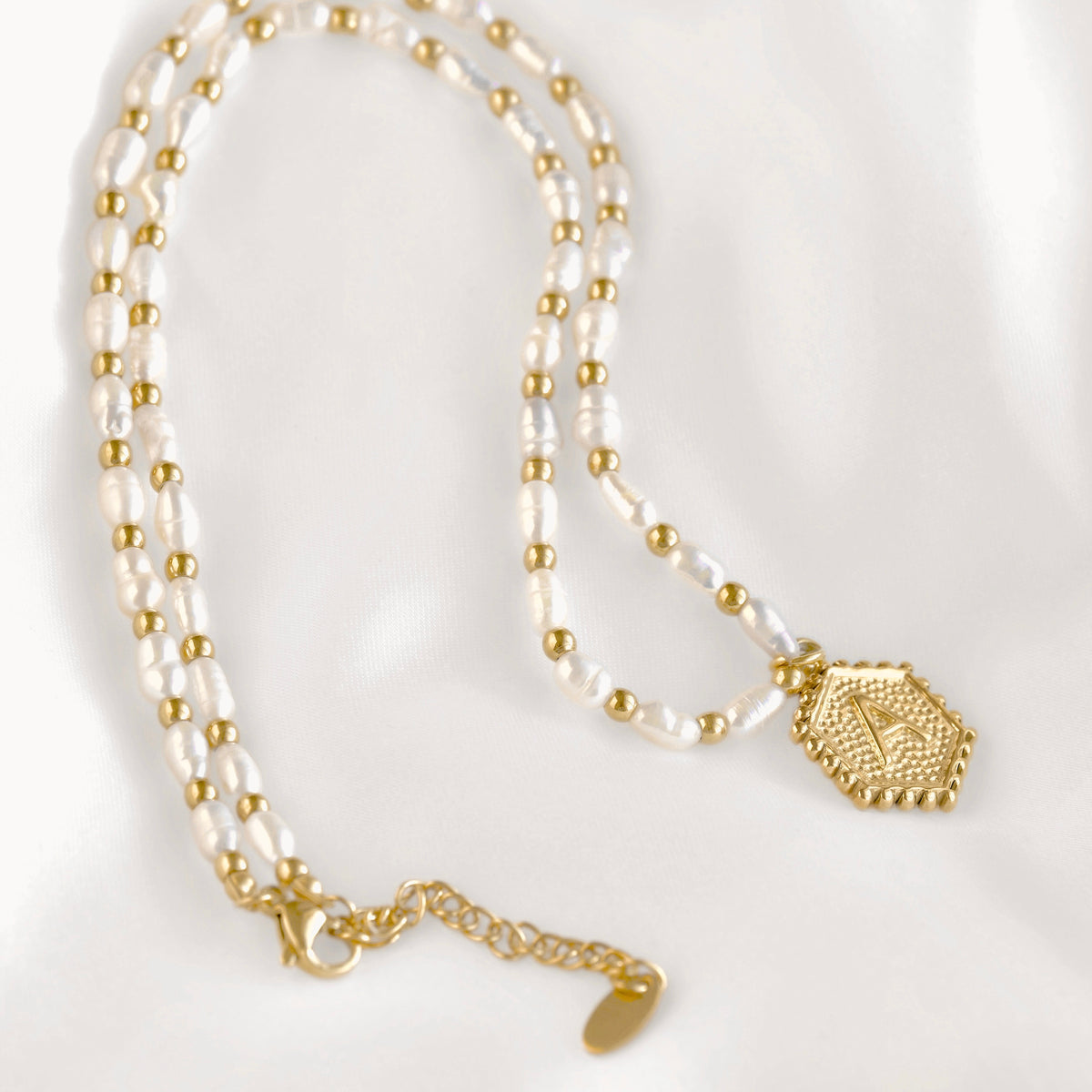 A double-strand necklace featuring white freshwater pearls and small gold beads, with a textured gold hexagonal pendant and gold chain extender, displayed on a white fabric background.