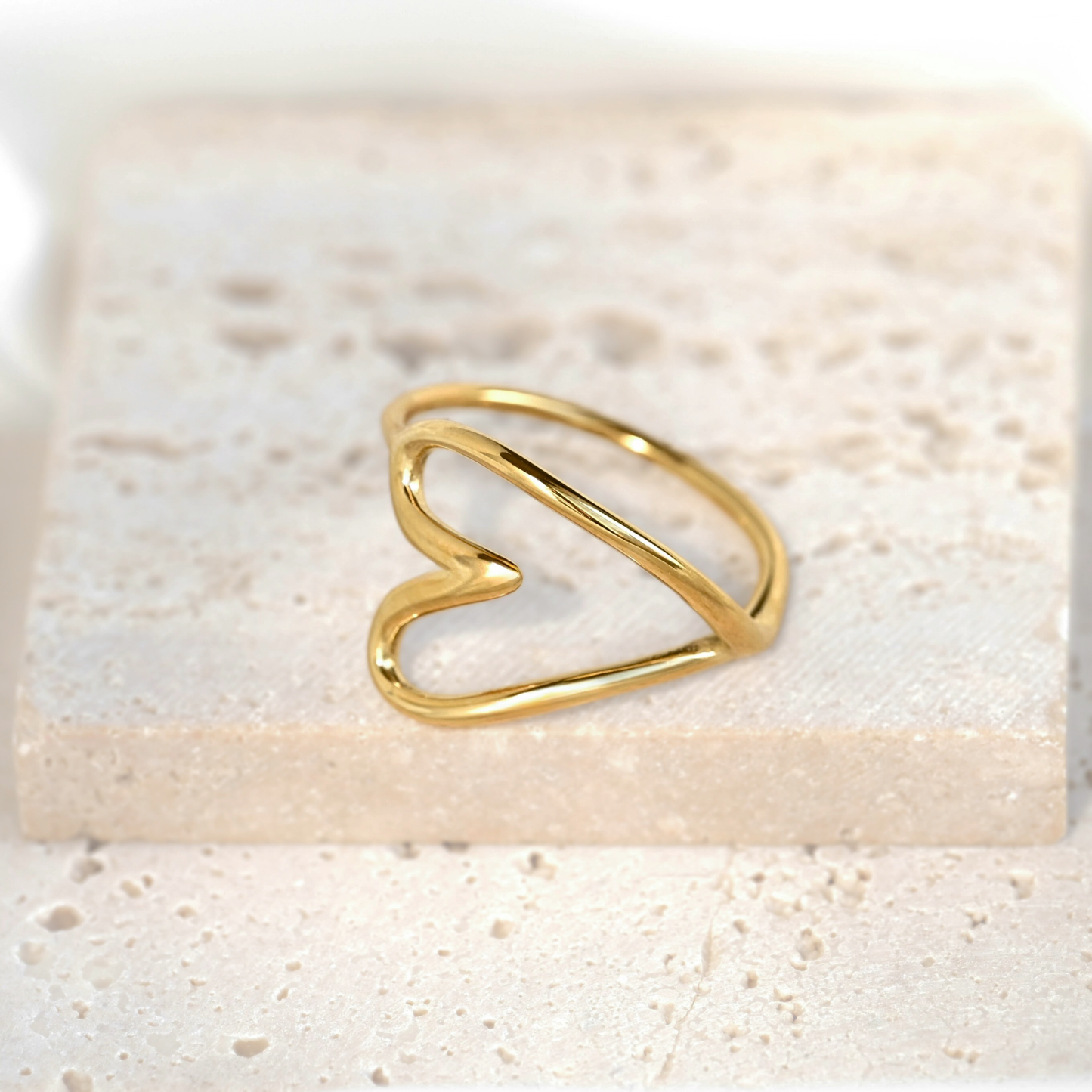A delicate gold wire ring shaped like an open heart, resting on a textured beige stone tile.