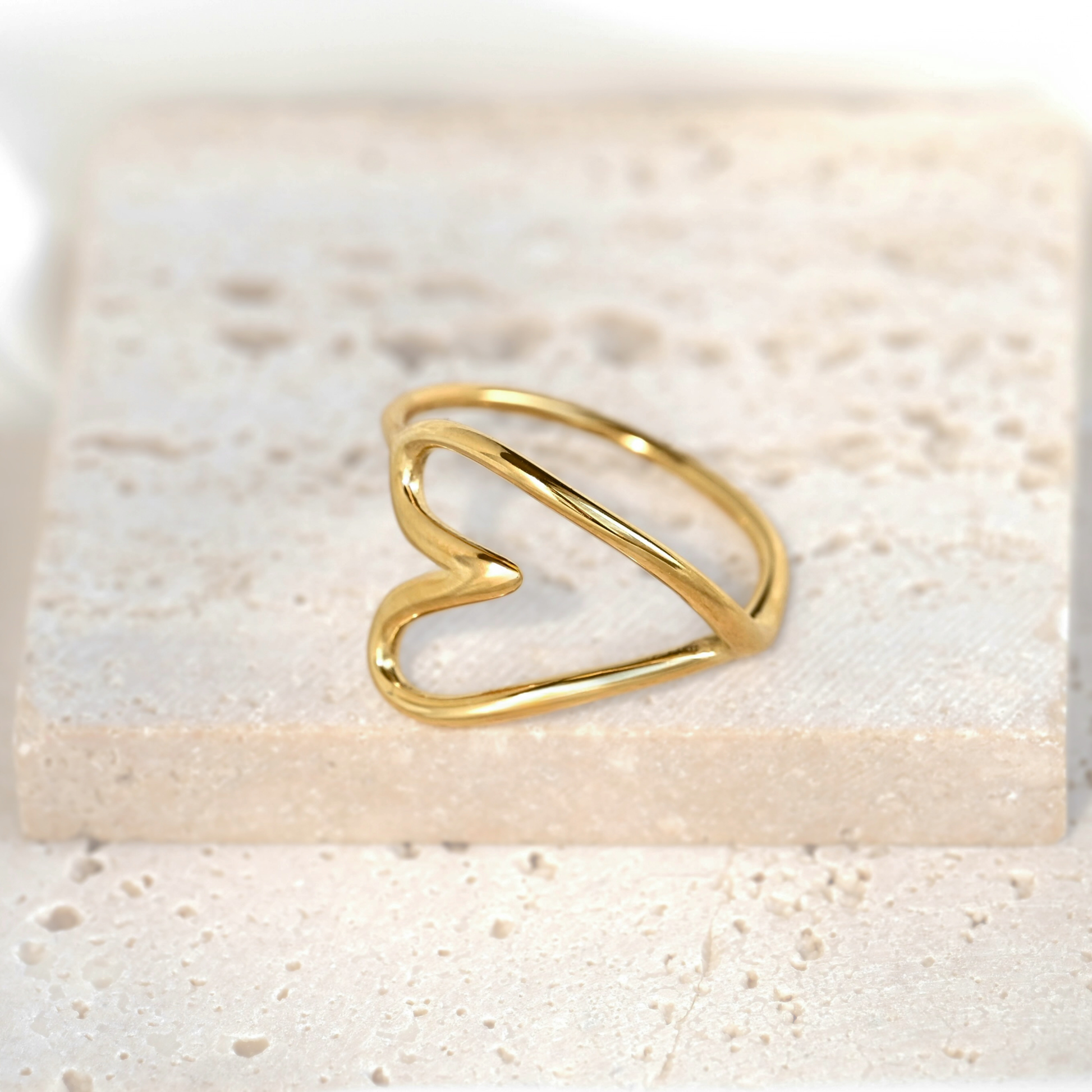 A delicate gold wire ring shaped like an open heart, resting on a textured beige stone tile.