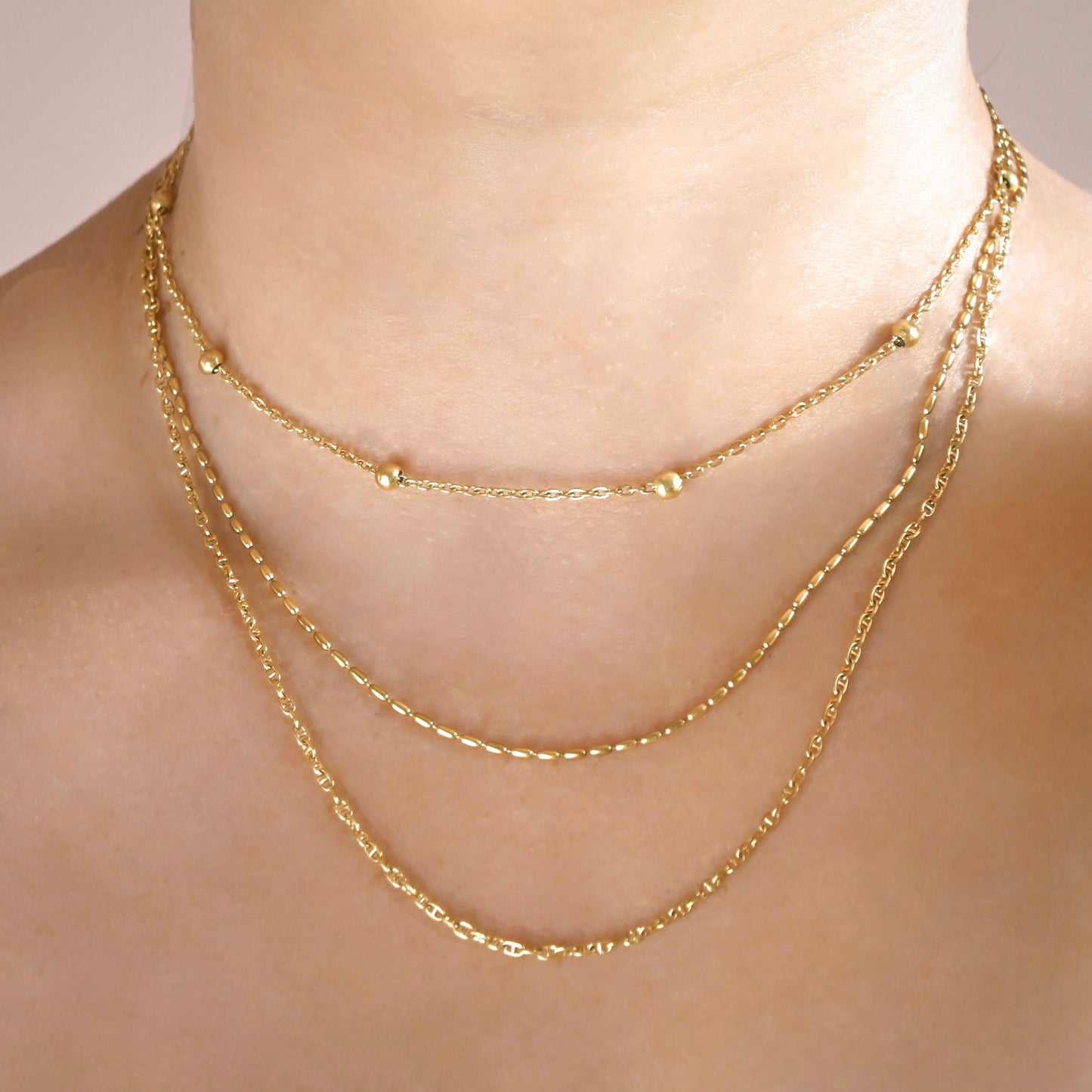 A close-up of a model's neck and collarbone wearing a three-layered gold necklace set, featuring delicate chains with small bead accents.