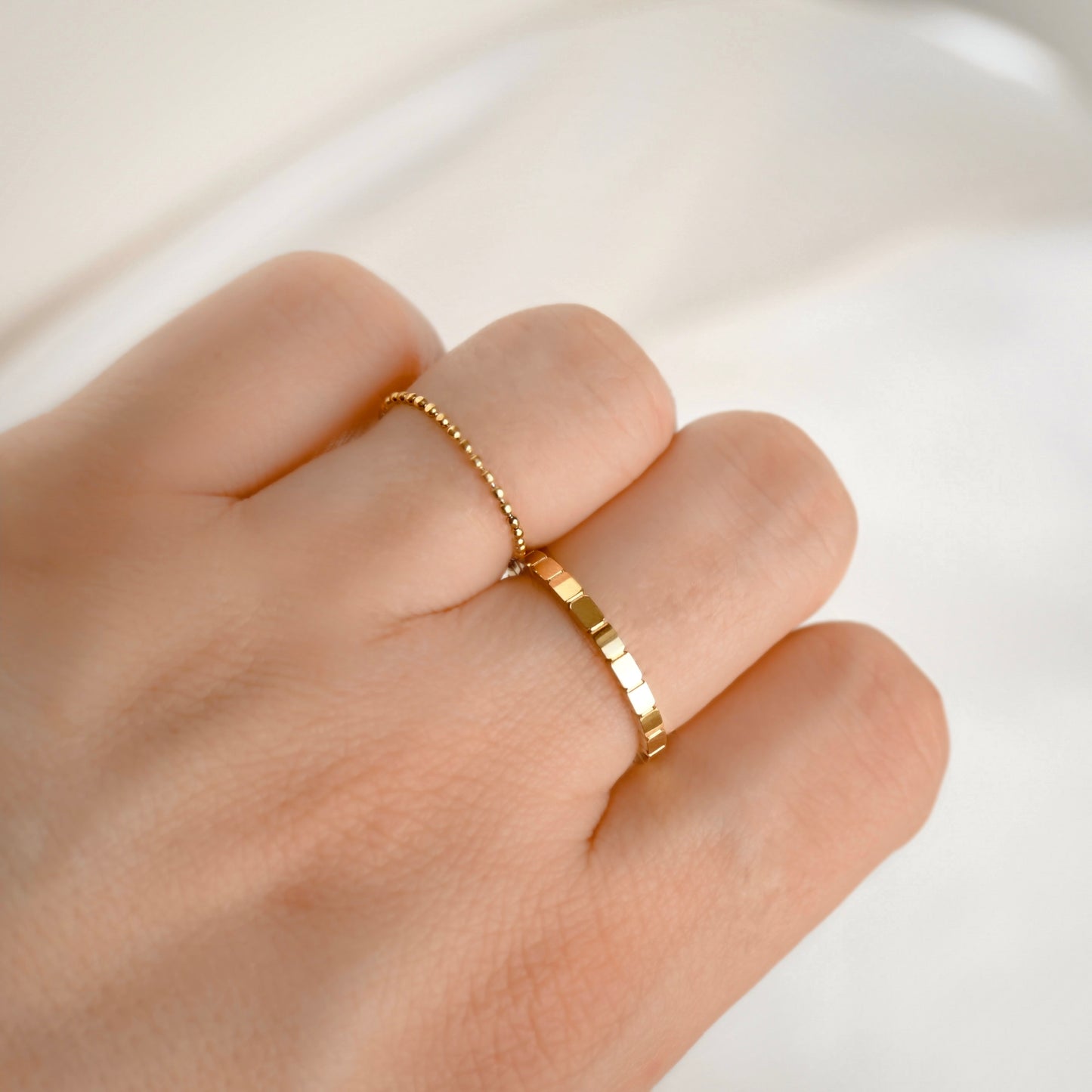A close-up of a hand wearing two delicate gold rings, including a thin beaded ring and a textured band ring.
