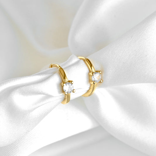 Two gold huggie earrings with clear gemstones displayed on a white satin ribbon.