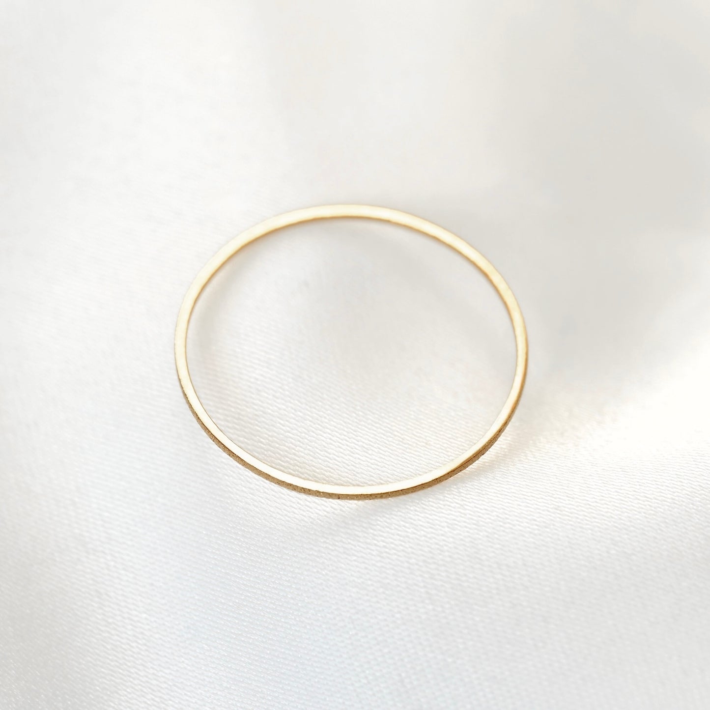 Top view of a minimalist, thin gold band ring with a textured finish with the top side smooth and high polish perfectly reflecting light, on a soft, white textured fabric background.