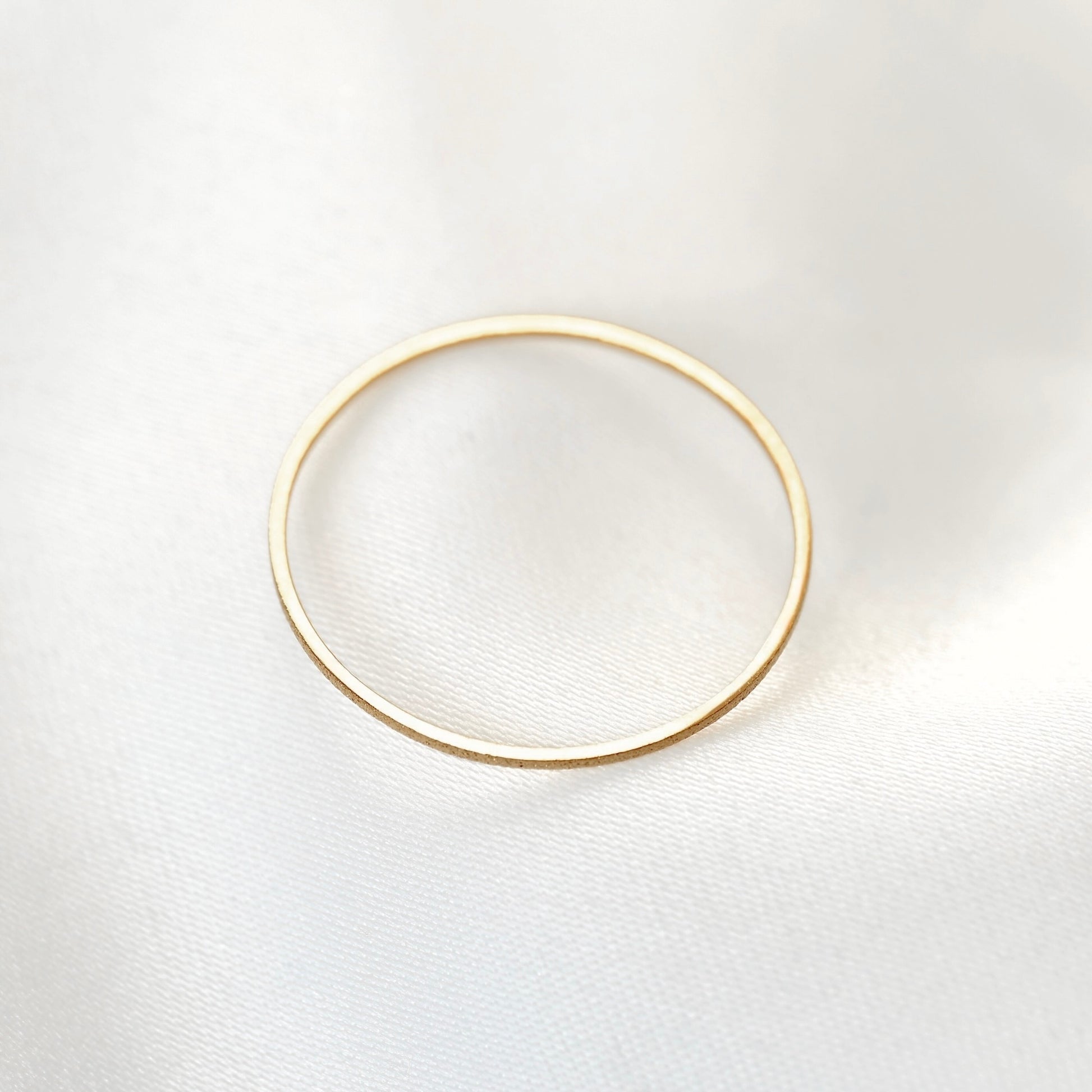Top view of a minimalist, thin gold band ring with a textured finish with the top side smooth and high polish perfectly reflecting light, on a soft, white textured fabric background.