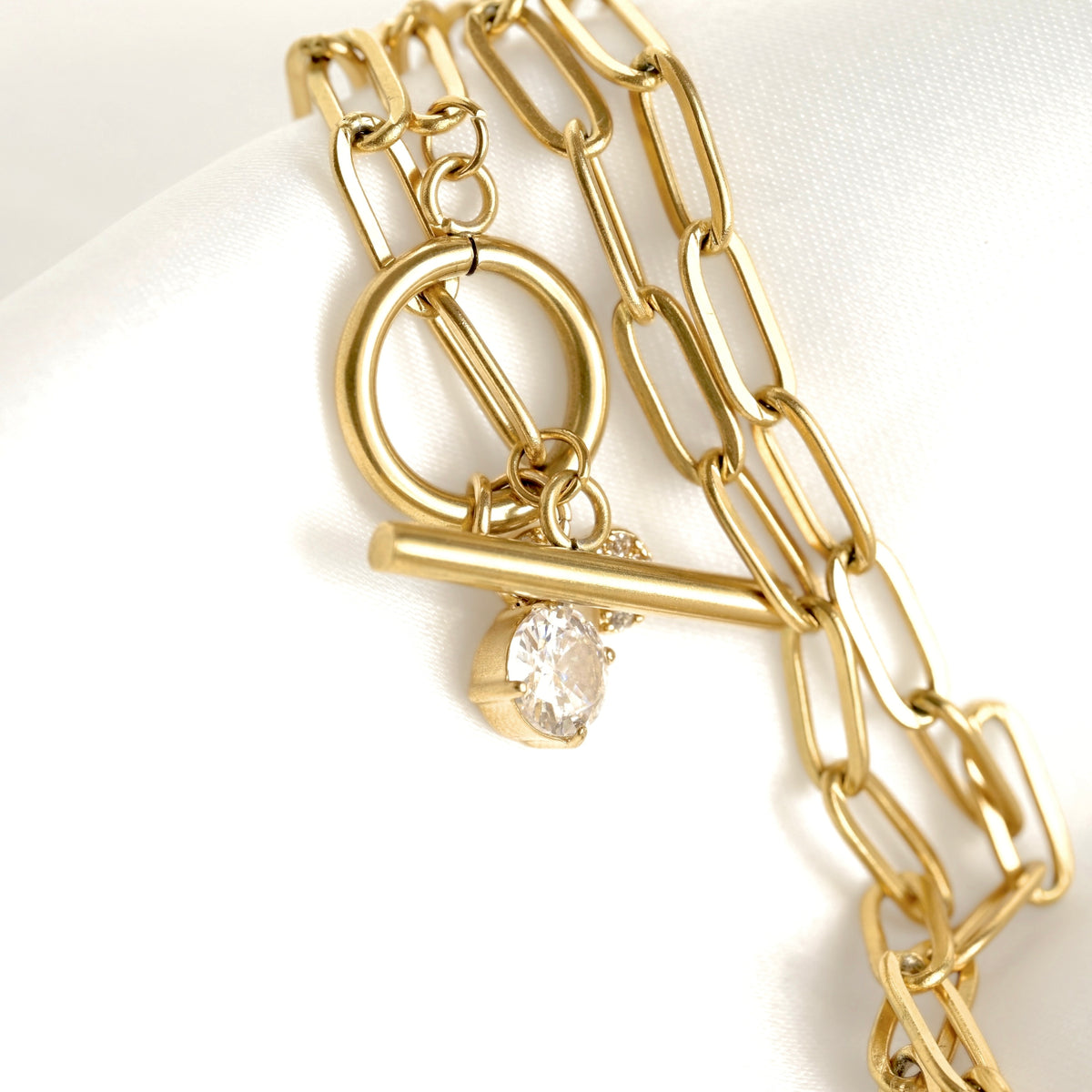 A gold chain necklace with a toggle clasp and a dangling clear round gemstone, displayed on a white satin surface.