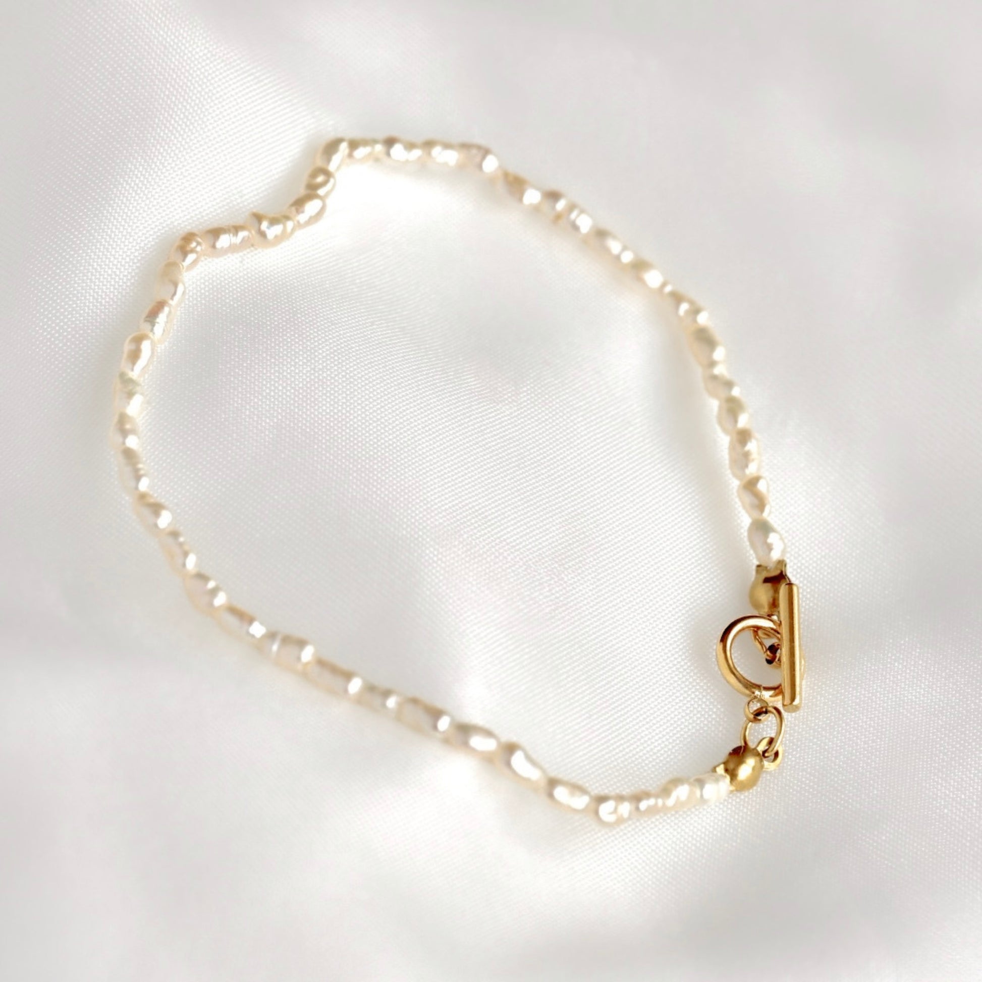 A delicate bracelet made of small irregular white  rice pearls linked with a gold-tone toggle clasp, displayed on a soft white satin fabric background.