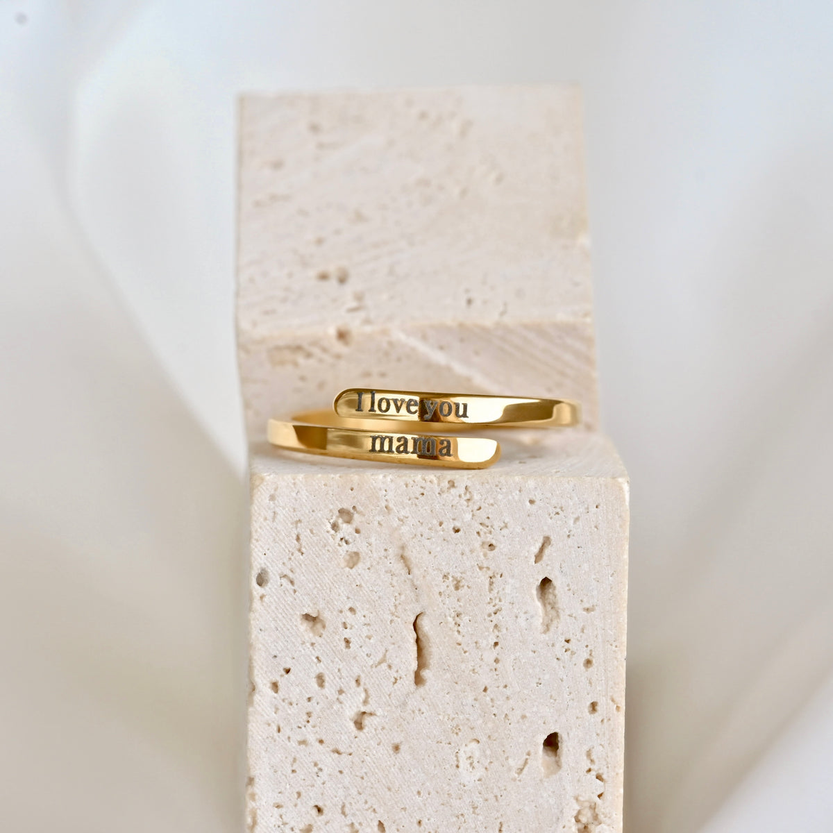A gold open cuff ring engraved with 'I love you' on top and 'mama' on bottom end in black lettering, displayed on a beige stone block against a soft white background.