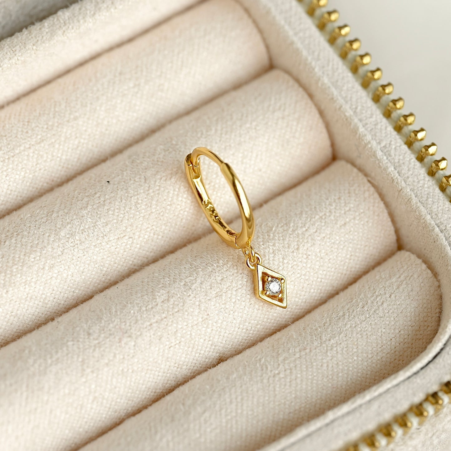 A gold huggie hoop ring with a small rhombus charm set with a clear CZ stone centerpiece, displayed inside a cream-colored jewelry box.