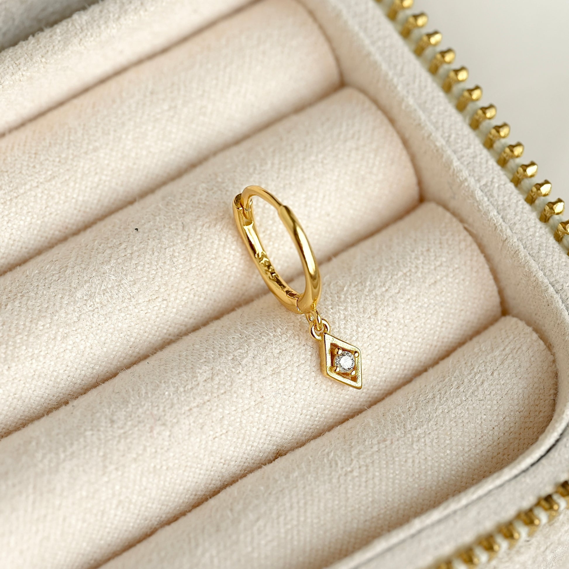 A gold huggie hoop ring with a small rhombus charm set with a clear CZ stone centerpiece, displayed inside a cream-colored jewelry box.