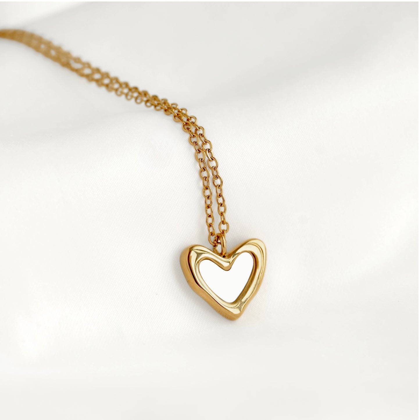 A gold heart-shaped pendant necklace with an open center, resting on a soft white fabric background.