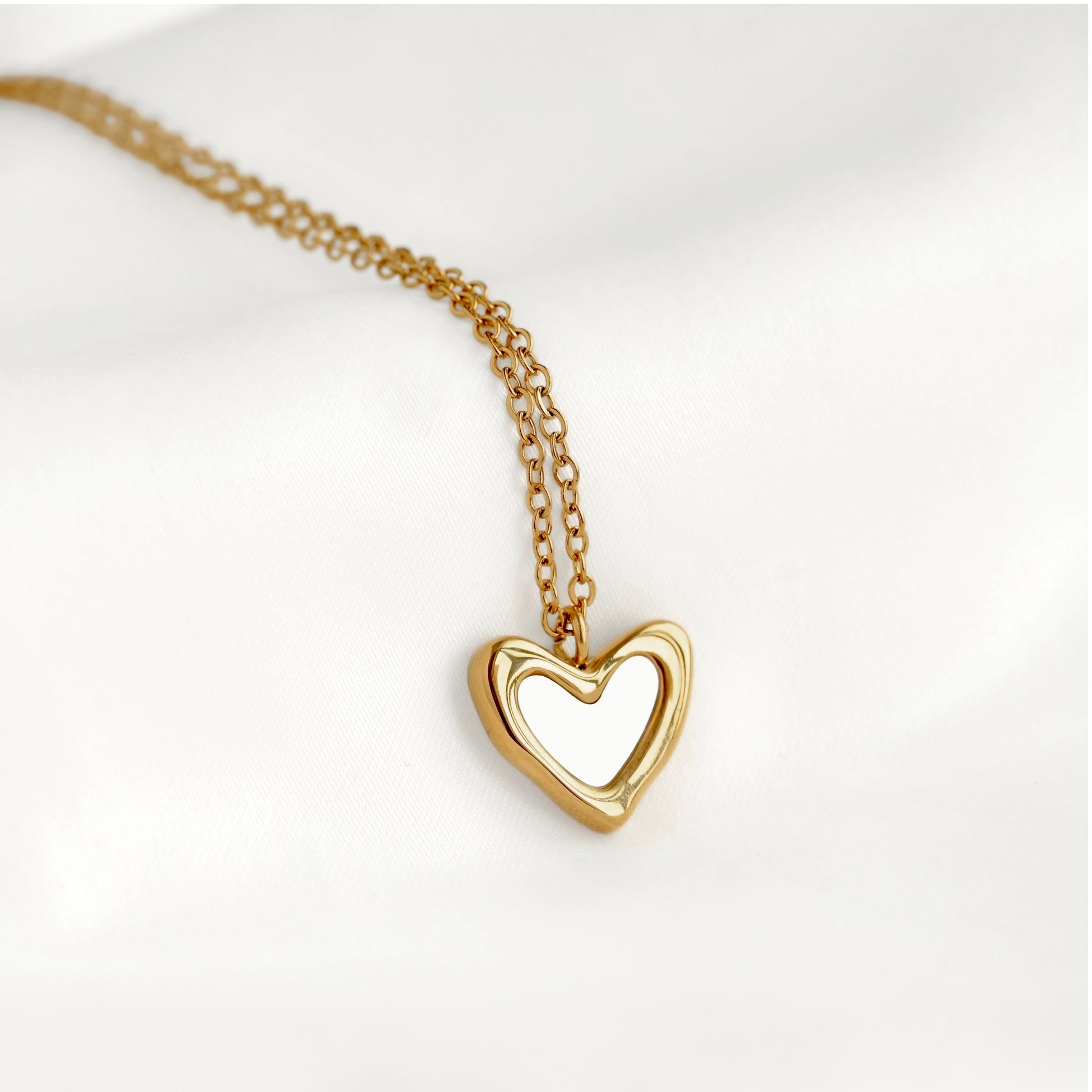 A gold heart-shaped pendant necklace with an open center, resting on a soft white fabric background.