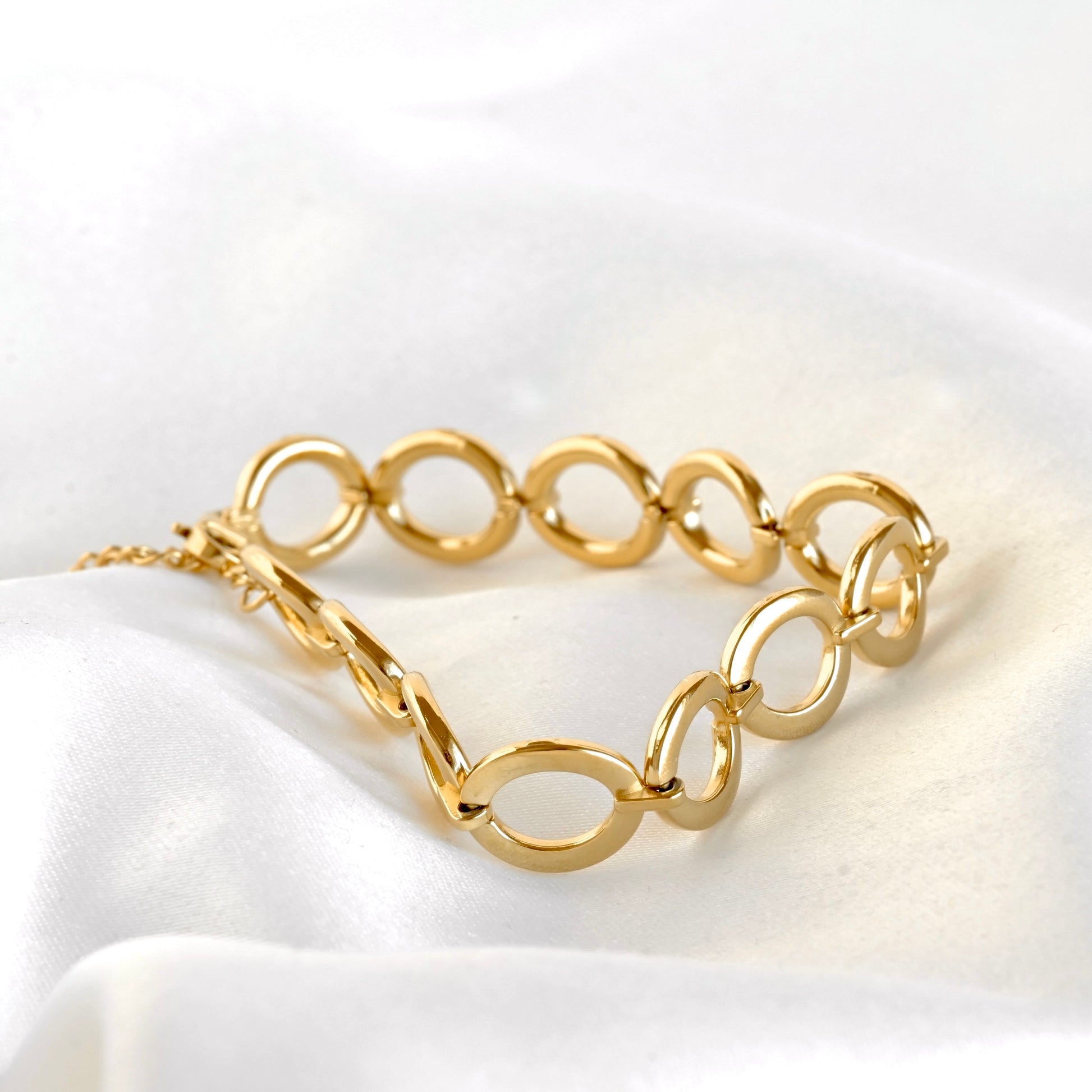 A gold chain bracelet with elongated oval links, displayed on a draped white satin fabric background.