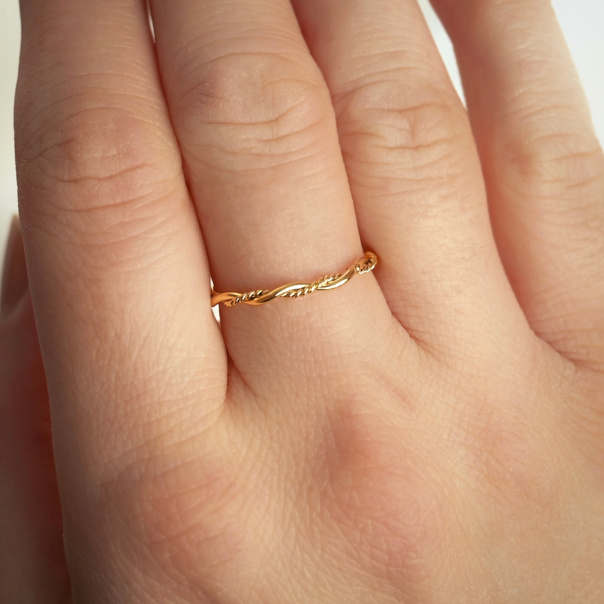 A delicate, thin gold ring with a twisted vine design worn on the ring finger of a hand.