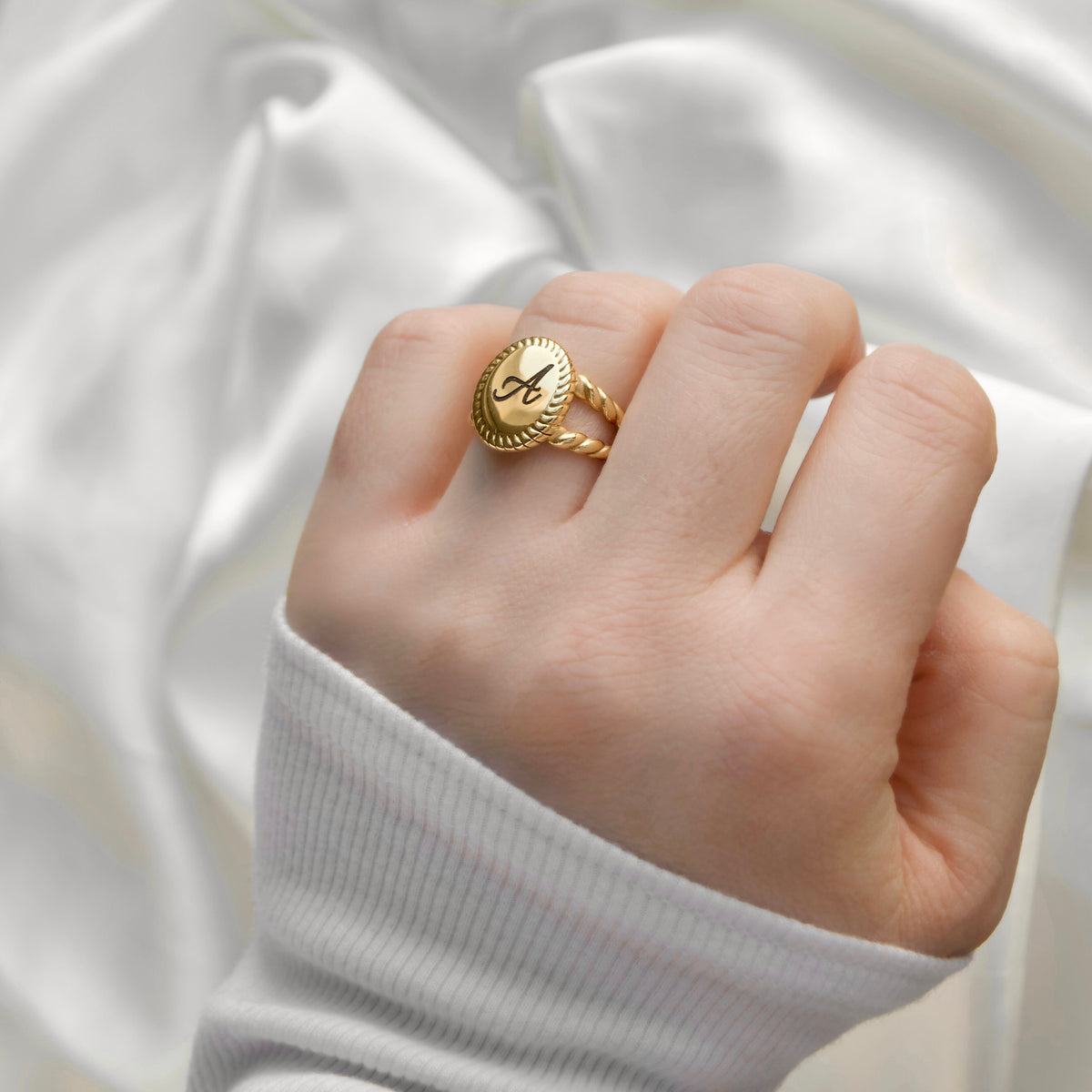 A close-up of a hand wearing a gold signet ring with a monogram 'M' on a draped white satin fabric background.