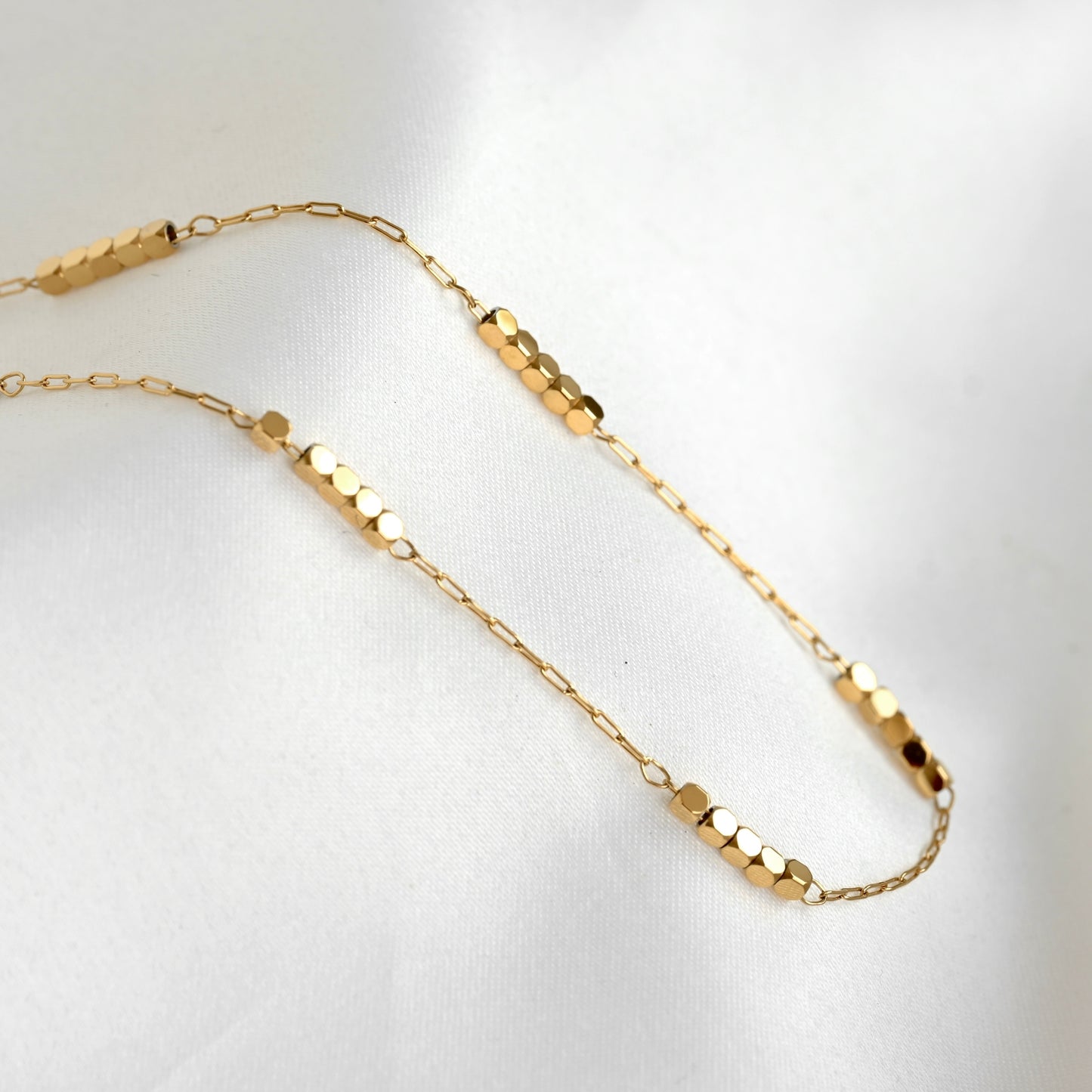 A delicate gold chain necklace featuring multiple segments of small cubic beads, displayed on a soft white fabric background.