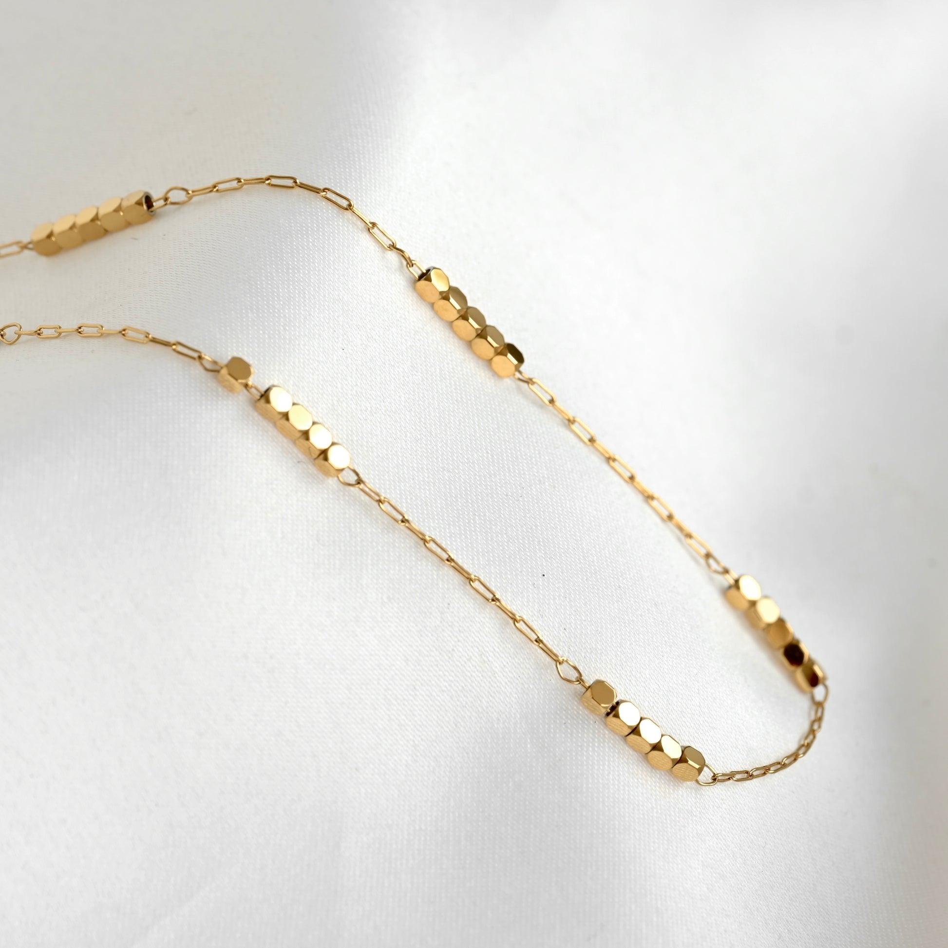 A delicate gold chain necklace featuring multiple segments of small cubic beads, displayed on a soft white fabric background.