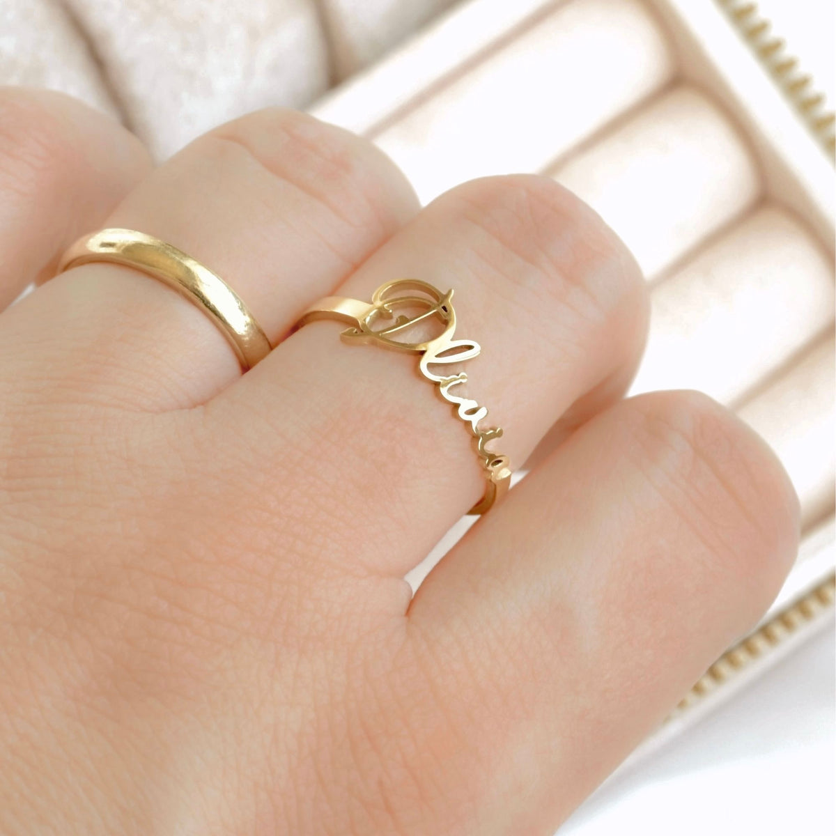 A close-up of a hand wearing a delicate gold ring with the name 'Olivia' written in cursive script, alongside a simple gold band, set against a soft-focus cream-colored jewelry box.