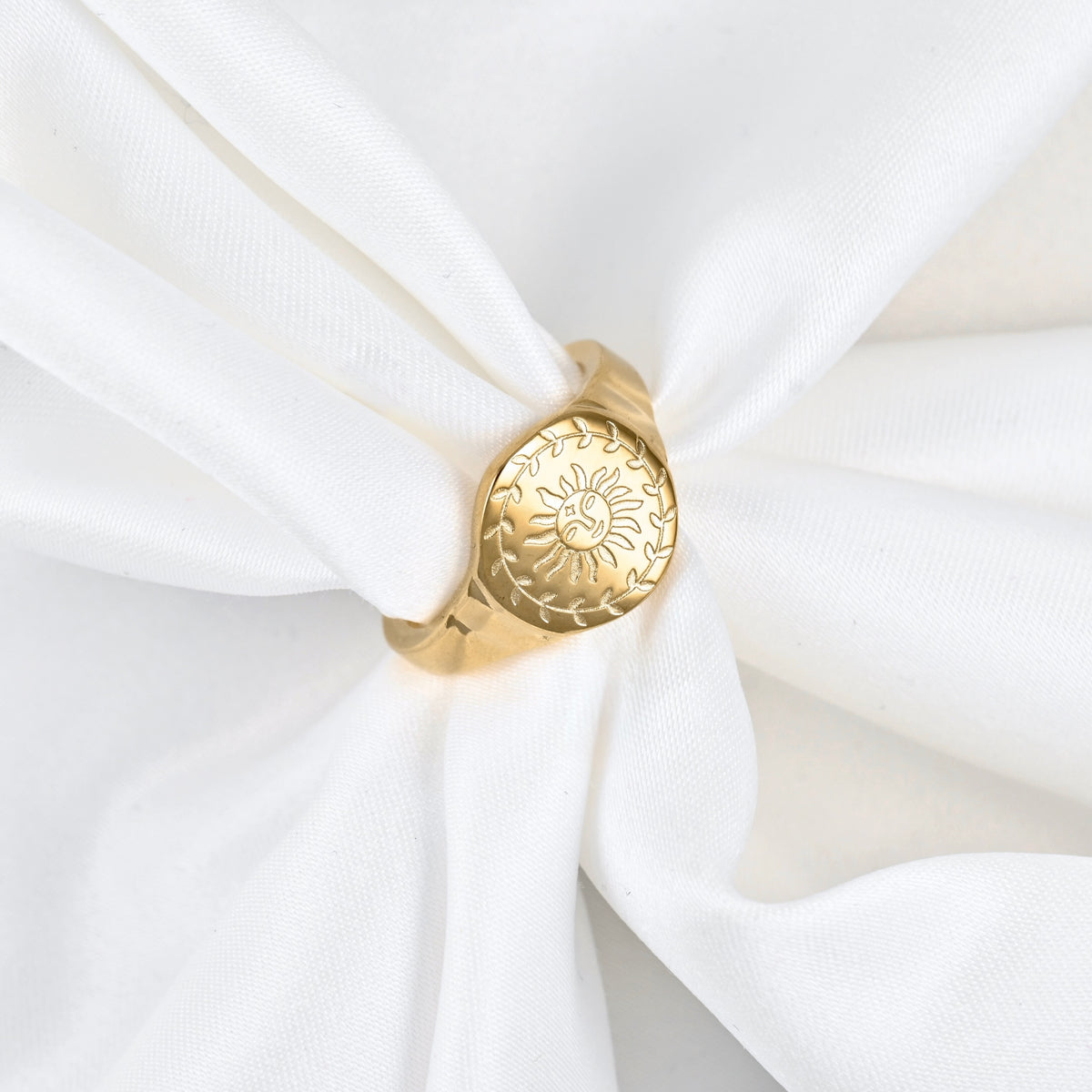 A gold signet ring with an engraved sun and leaf pattern, displayed on a white fabric napkin.