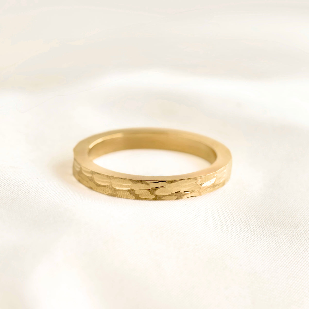 A gold band ring with a textured, hammered finish resting on a soft, white fabric background.