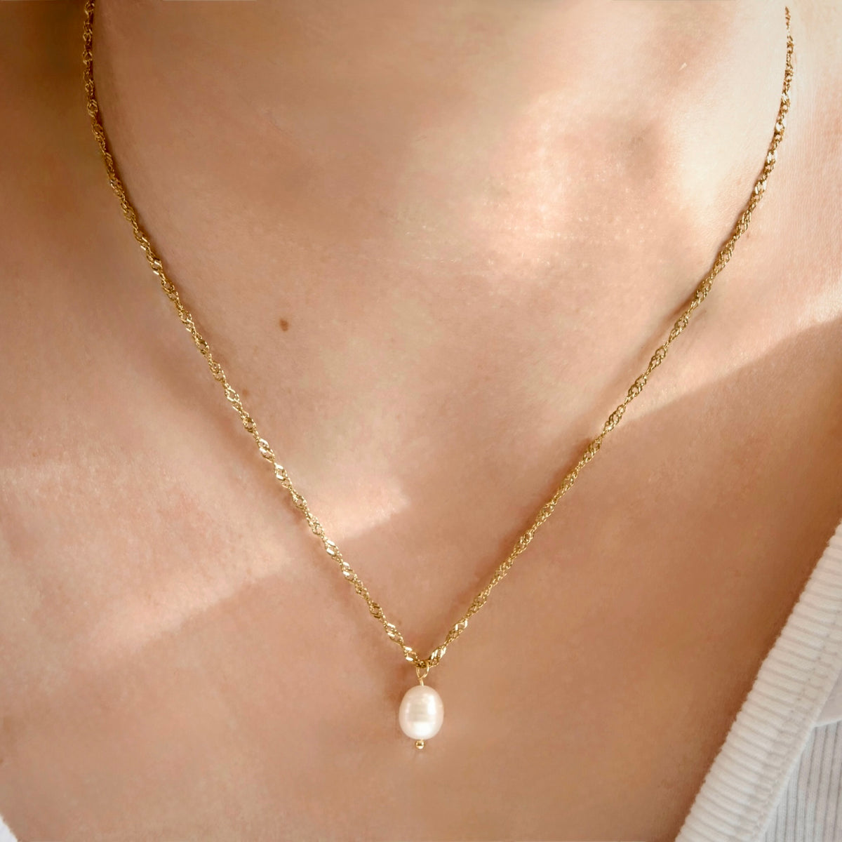 A delicate gold singapore chain necklace with a single small white pearl pendant, worn on a person's neck.