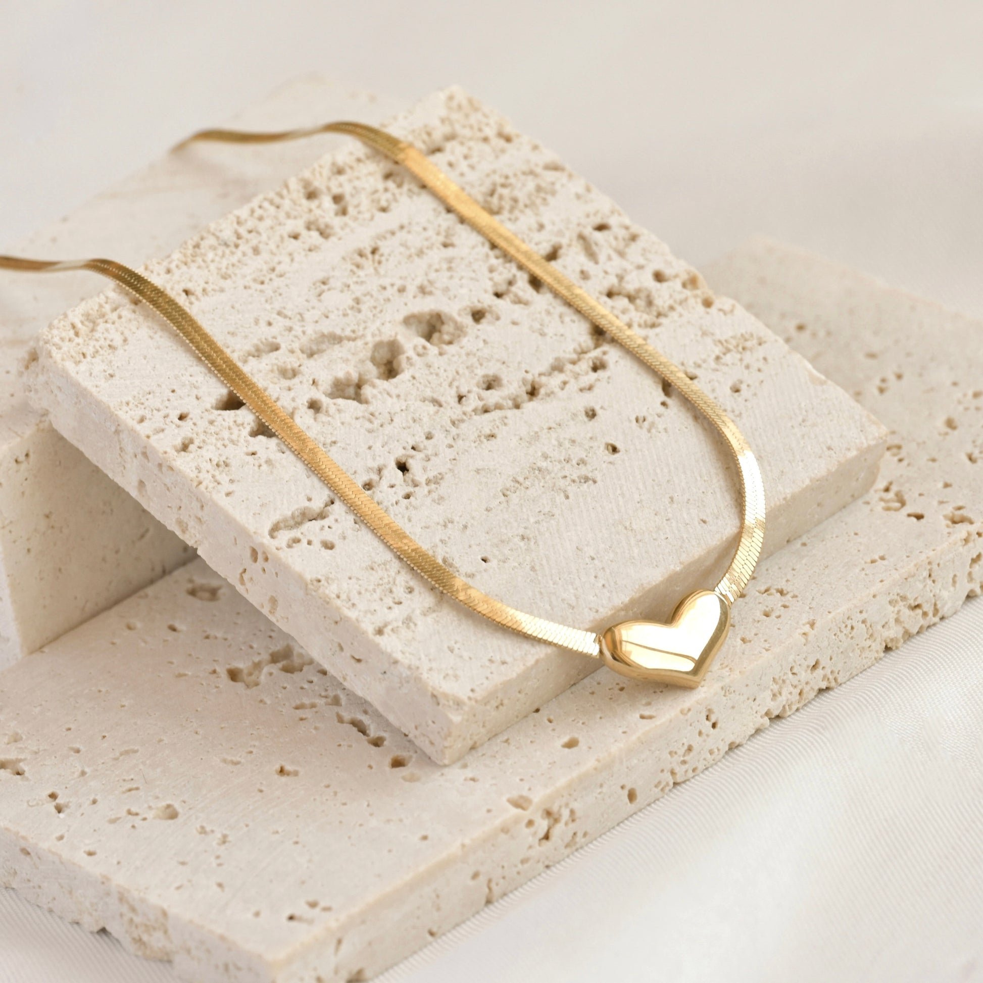 Gold herringbone necklace with a heart charm on a textured stone surface