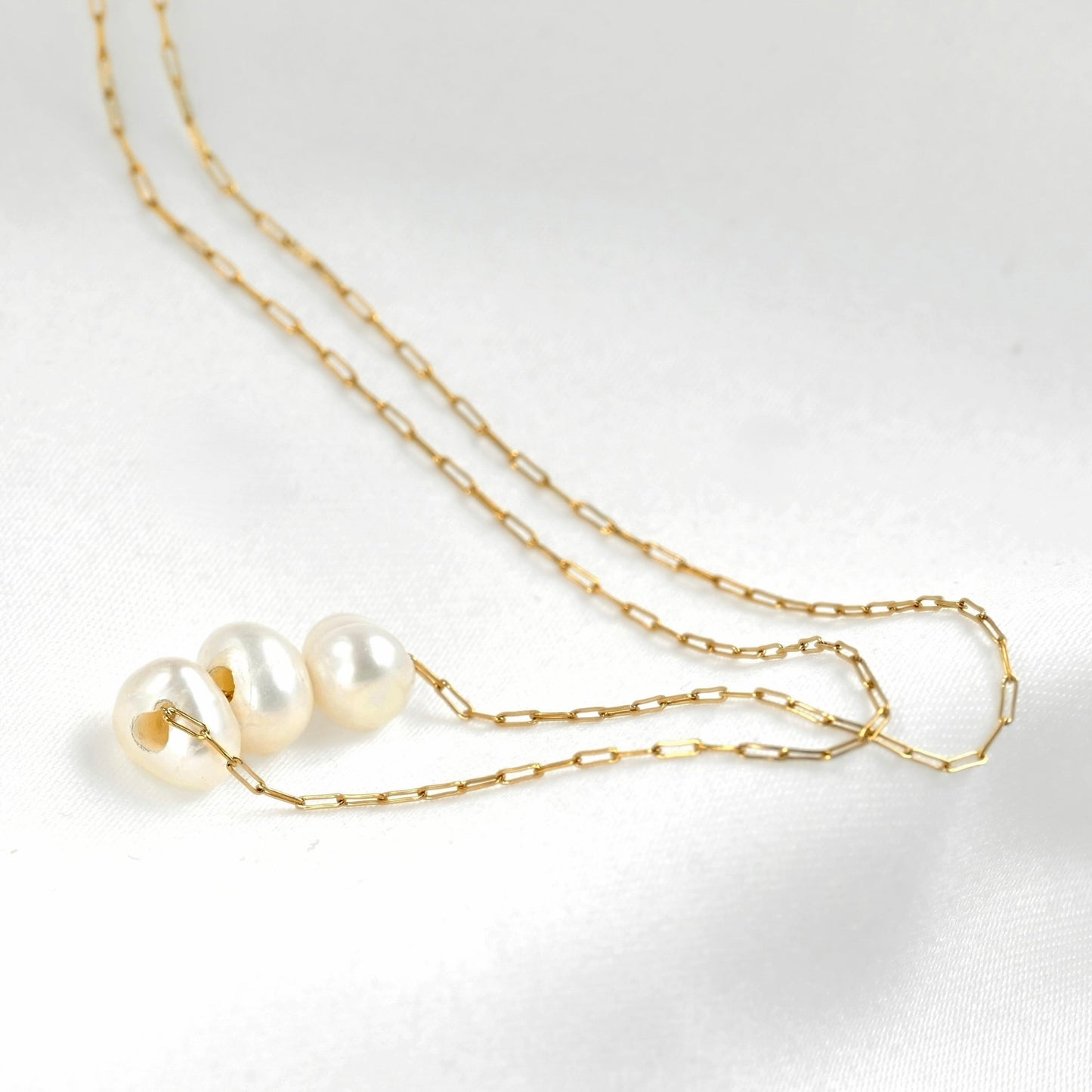A delicate gold chain necklace featuring three small, lustrous white pearls arranged in a row, displayed on a clean white fabric background.