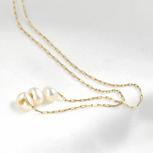 A delicate gold chain necklace featuring three small, lustrous white pearls arranged in a row, displayed on a clean white fabric background.