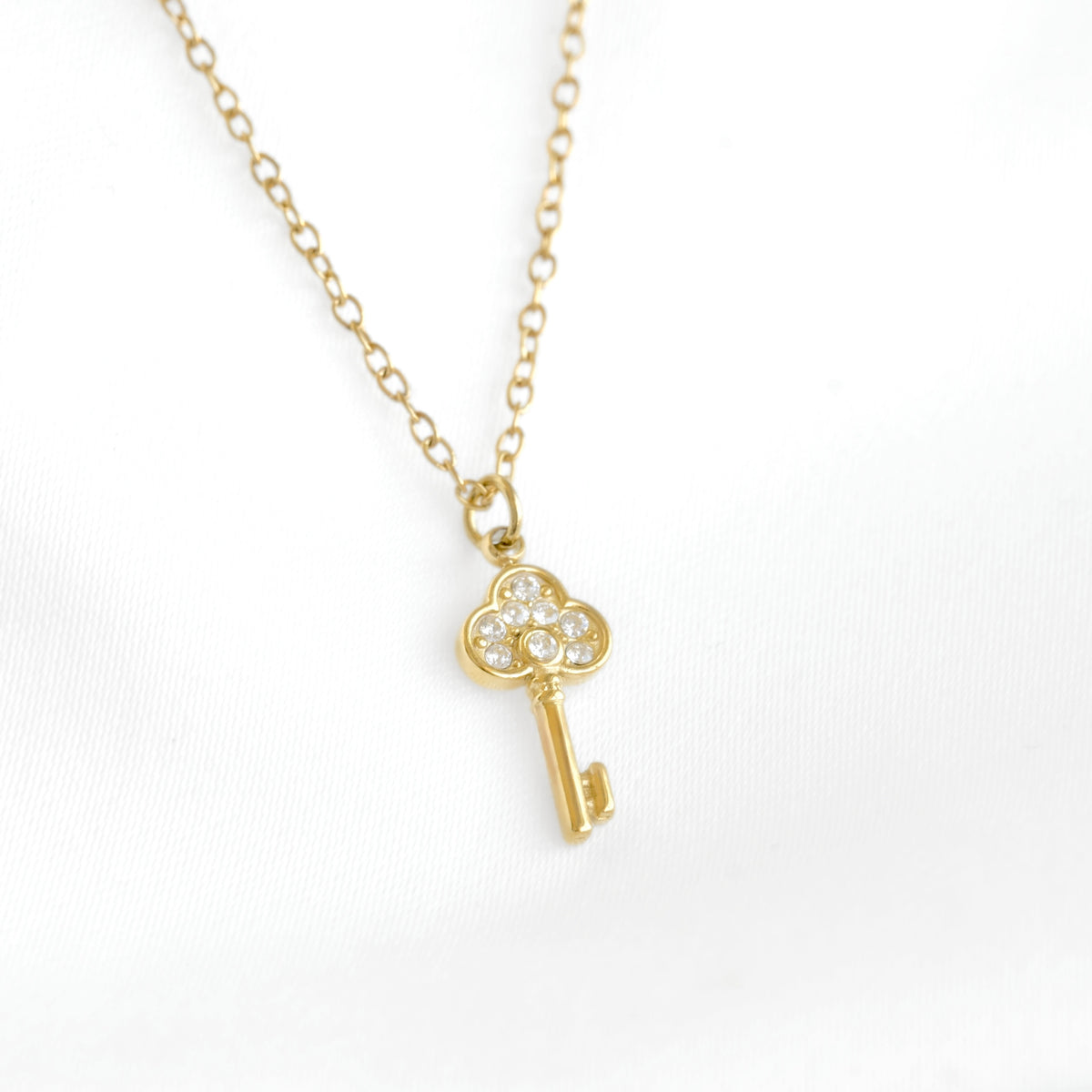 A gold key pendant necklace with a small cluster of clear stones on the key head, displayed on a white fabric background.