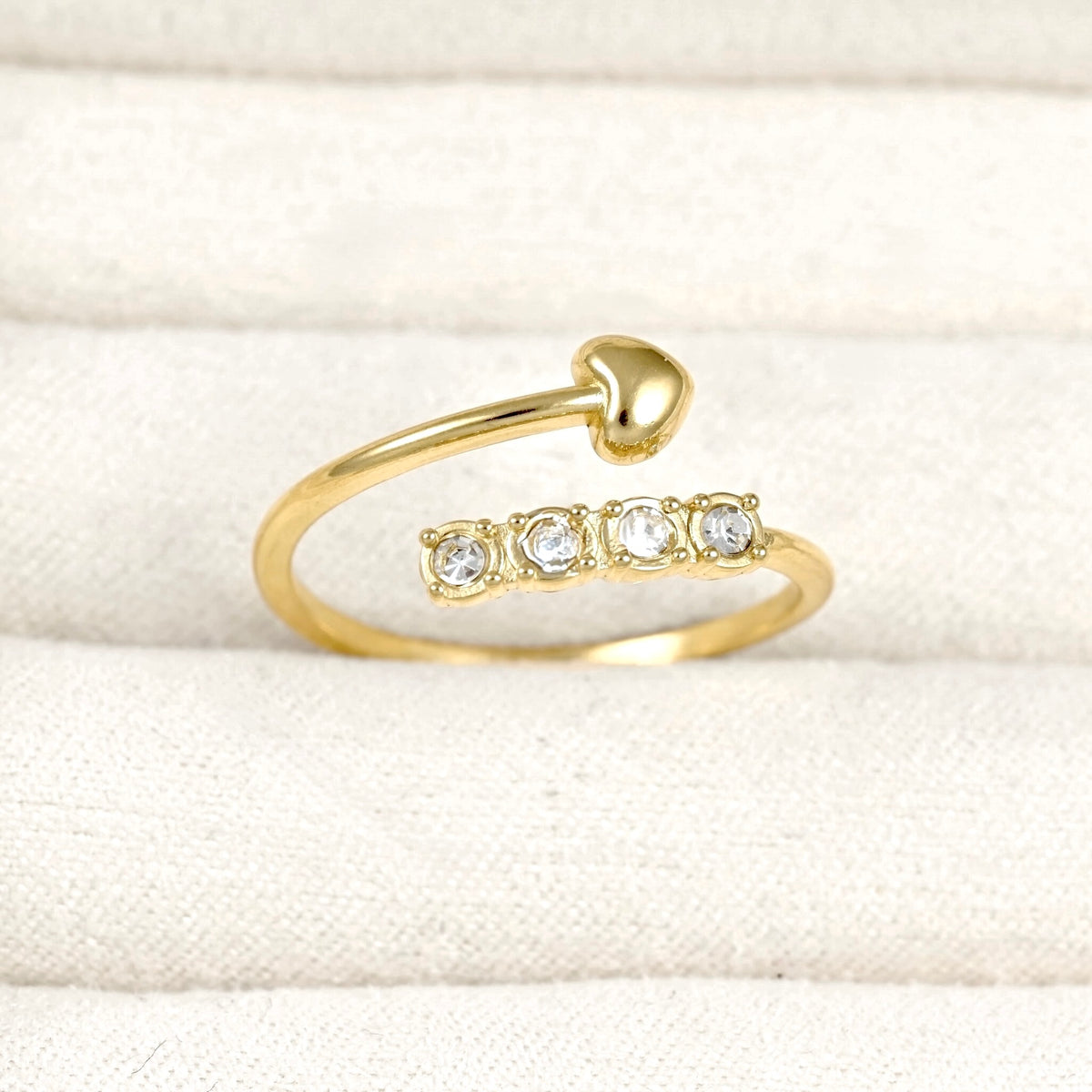 A gold open-band ring with a chevron shaped heart on top and four clear round gemstones on bottom, displayed on a textured cream fabric background.