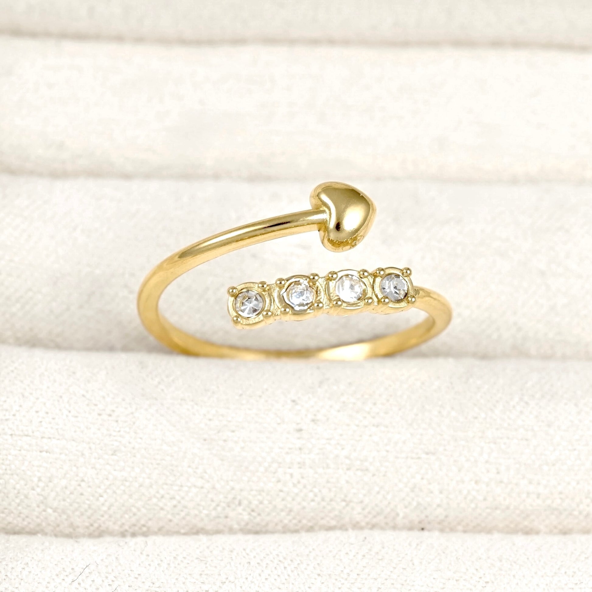 A gold open-band ring with a chevron shaped heart on top and four clear round gemstones on bottom, displayed on a textured cream fabric background.
