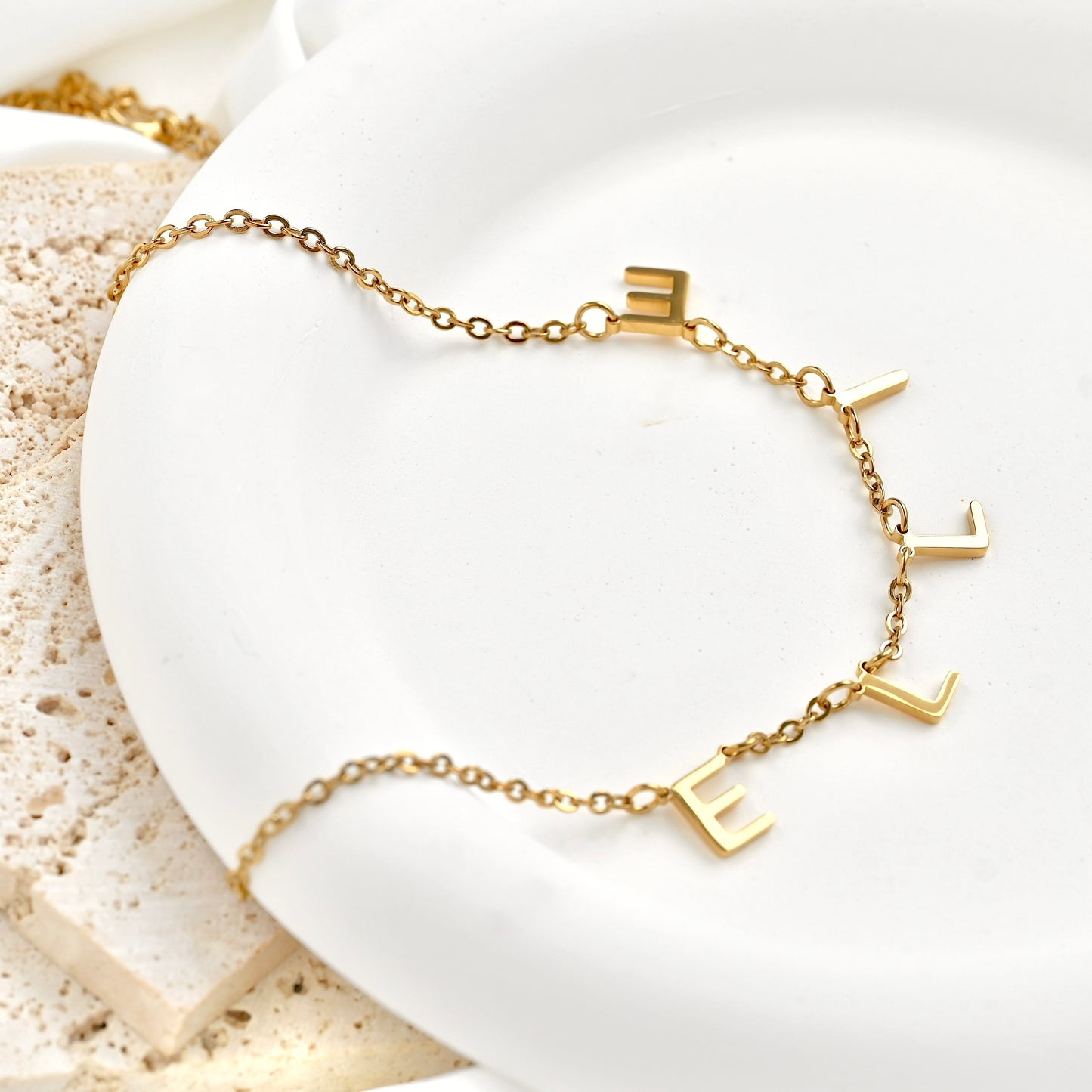 A delicate gold chain necklace with five raised letter charms spelling 'ELLIE', displayed on a white ceramic dish with a textured stone prop.