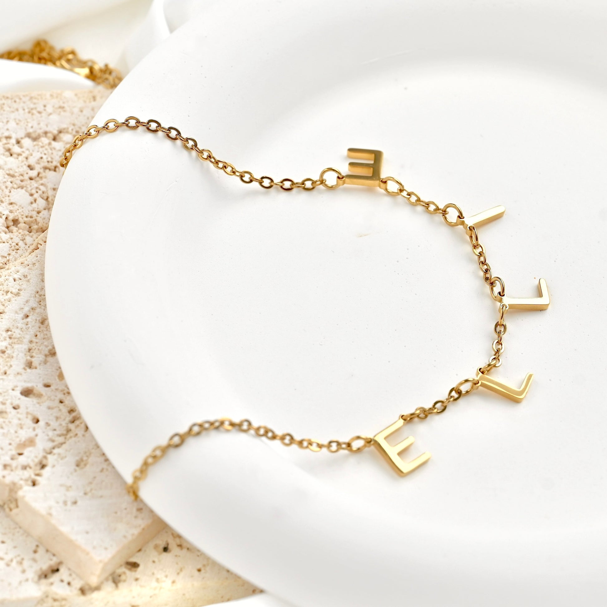 A delicate gold chain necklace with five raised letter charms spelling 'ELLIE', displayed on a white ceramic dish with a textured stone prop.