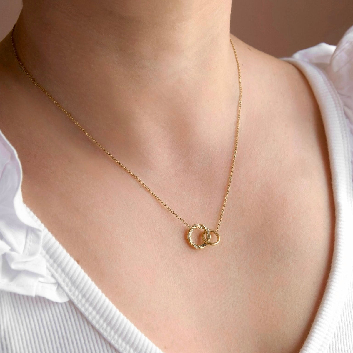 A close-up of a model wearing a delicate gold chain necklace with two interlocking open circle pendants, styled with a white ruffled-shoulder top.