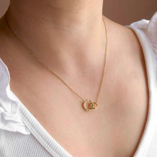 A close-up of a model wearing a delicate gold chain necklace with two interlocking open circle pendants, styled with a white ruffled-shoulder top.