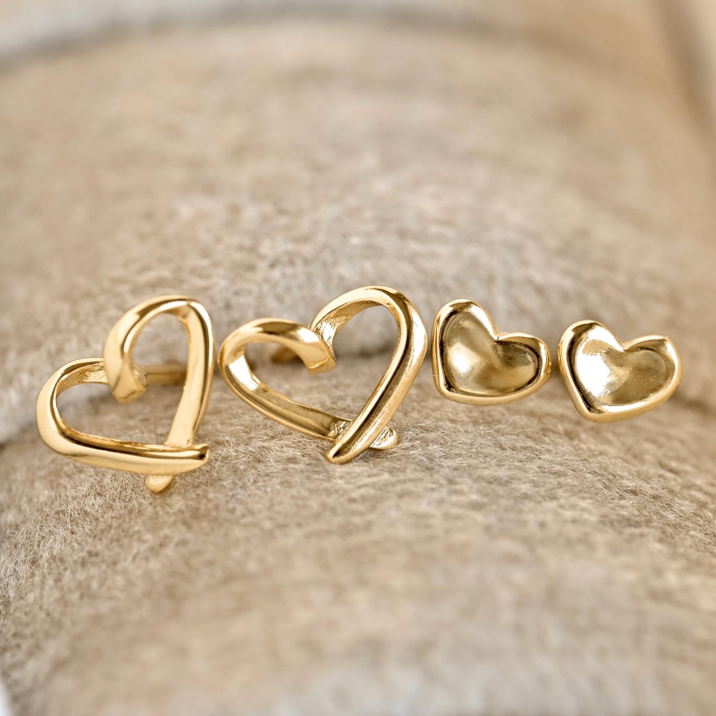 Four gold heart-shaped stud earrings arranged in ascending sizes on a textured beige surface.