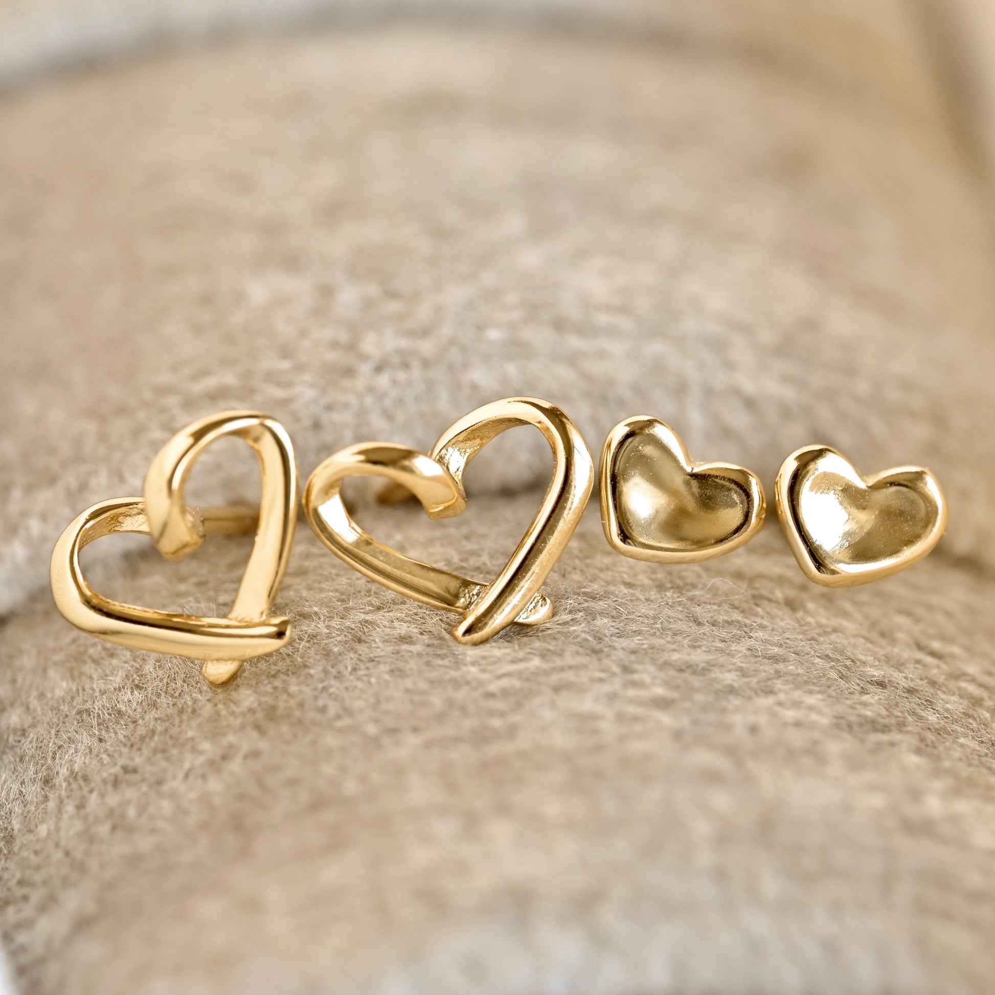 Four gold heart-shaped stud earrings arranged in ascending sizes on a textured beige surface.