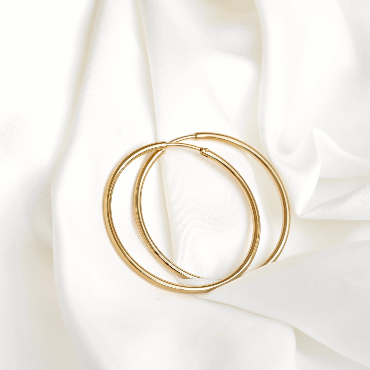 A pair of thin gold hoop earrings with endless closure resting on draped white fabric.