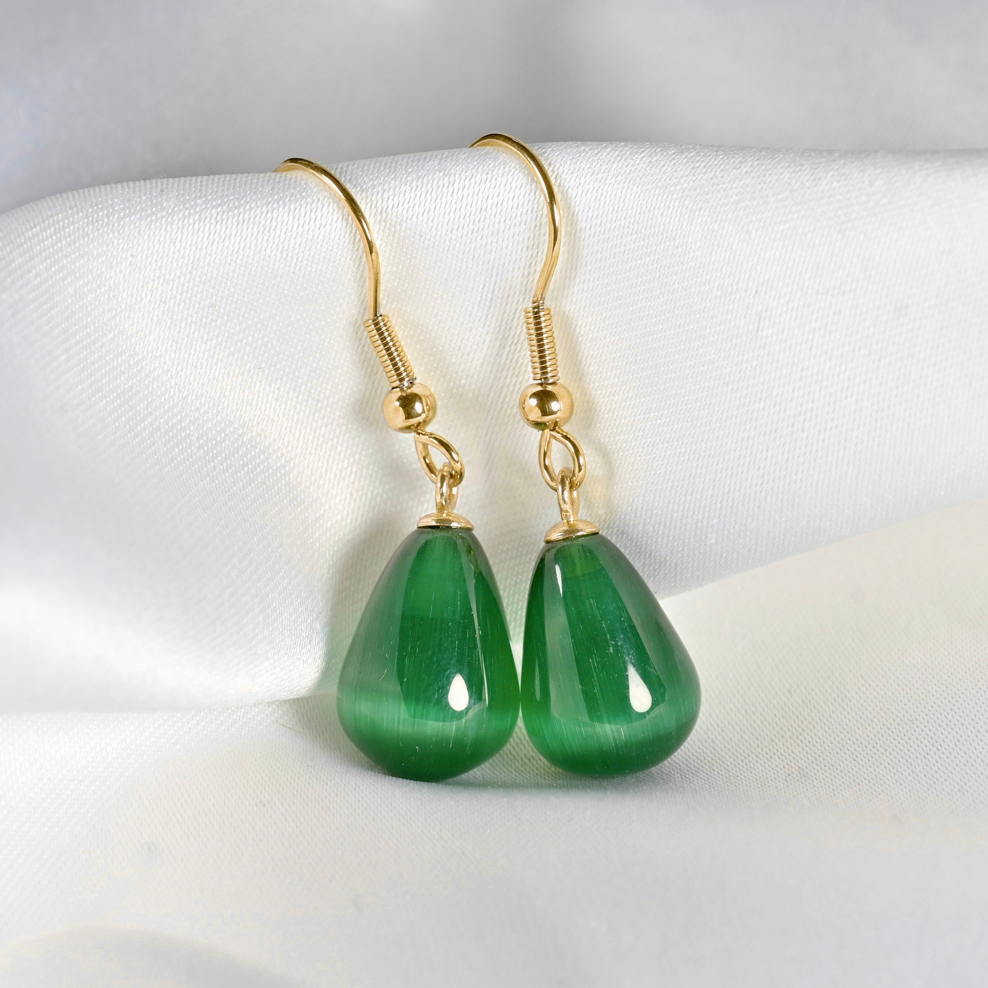 A pair of gold-tone dangle earrings with polished green teardrop-shaped stone drops, displayed on a soft white fabric surface.
