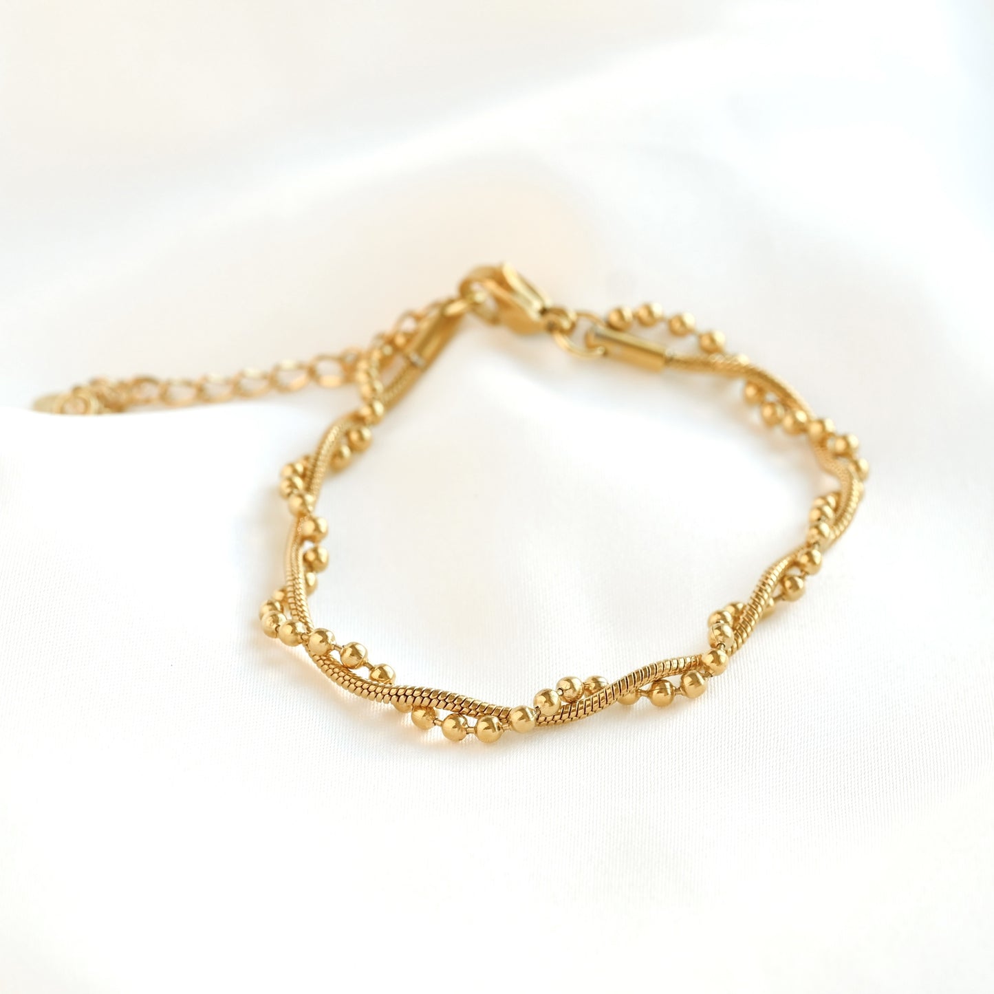 A delicate entwined gold chain bracelet with a wavy, beaded design, displayed on a soft white fabric background.