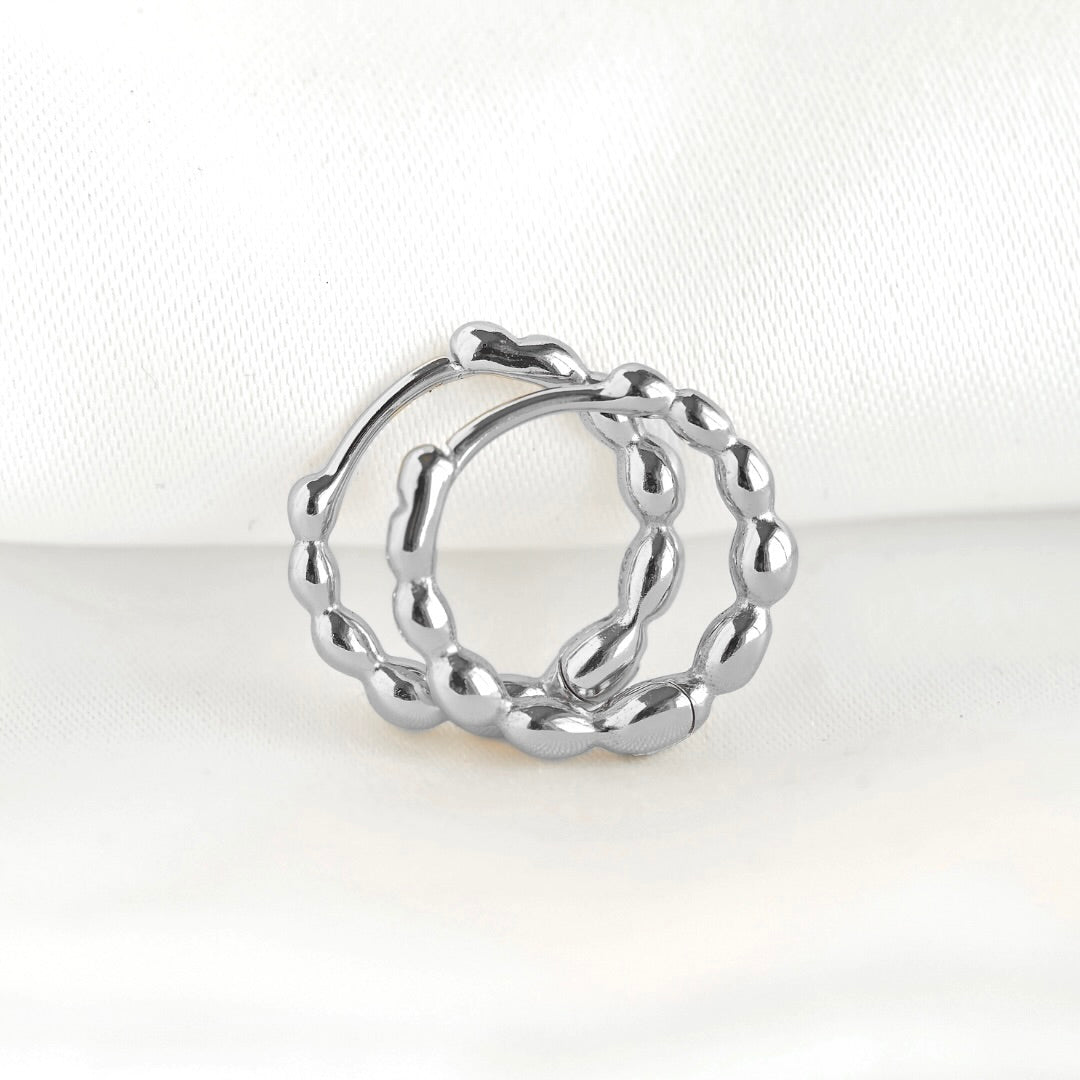 A silver-toned beaded hoop earring with a textured, beaded, and wavy design, displayed on a soft white fabric background.