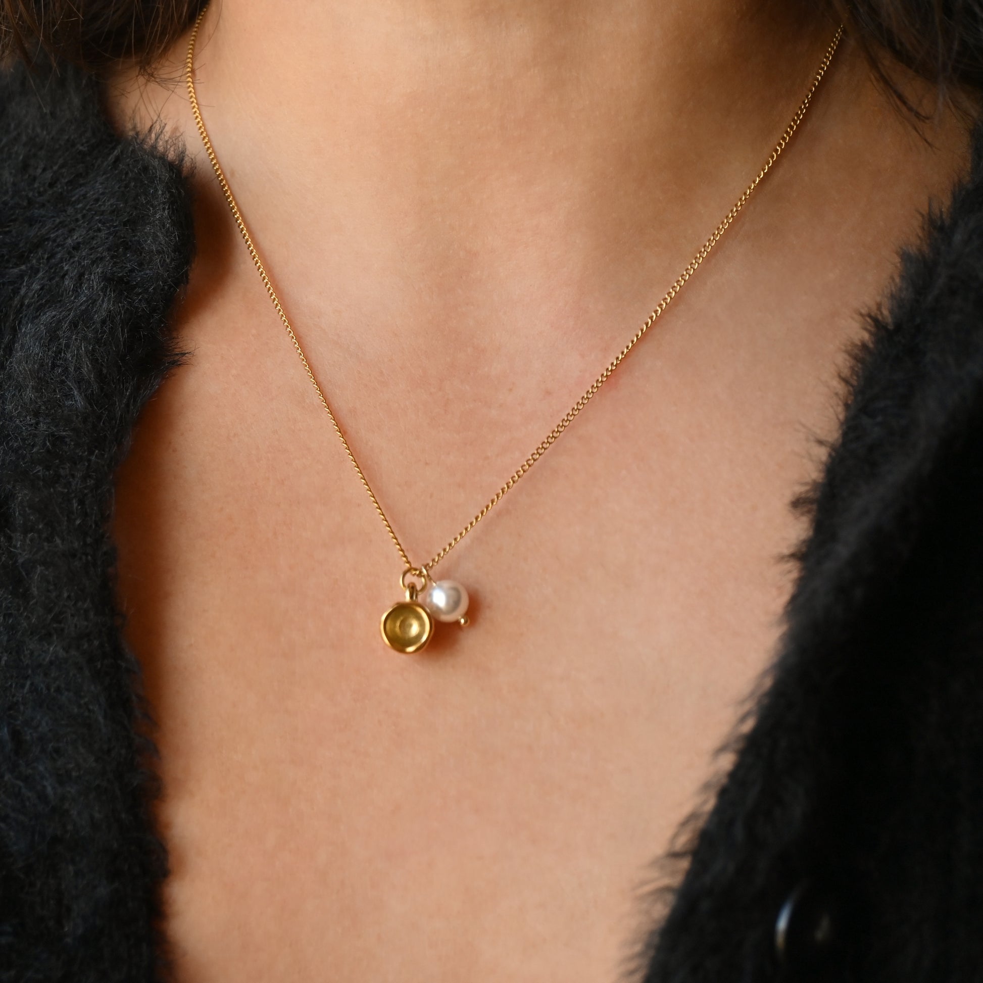 A close-up of a person wearing a delicate gold chain necklace with a double pendant design: one polished gold mug and one small white pearl, styled with a black fuzzy jacket.